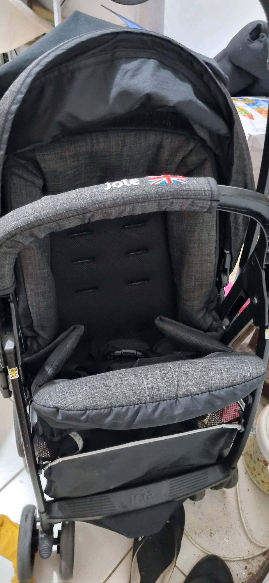 This image shows a close-up view of a dark gray Joie brand stroller. The stroller seat appears padded and has a mesh insert. A Union Jack flag is embroidered on the stroller's handle. The stroller is positioned indoors, on a tiled floor that is partially visible in the foreground. In the background, other objects are blurred, suggesting an indoor environment. The scene is likely in Denpasar, Indonesia, as indicated by the prompt. There are no people visible in the image.