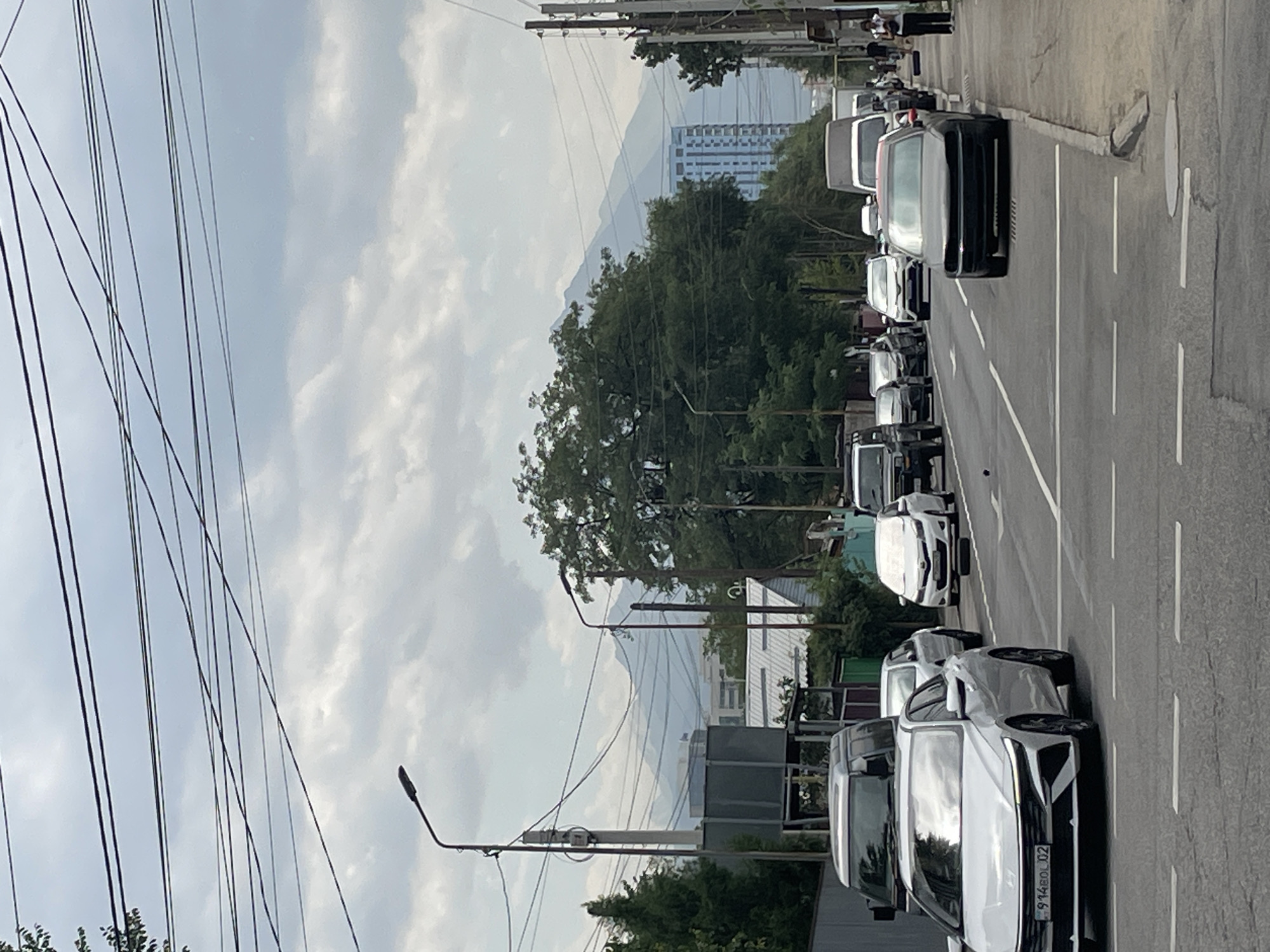 The image depicts a street scene in Almaty, Kazakhstan, with a view of the Tien Shan mountains in the background. The foreground shows a multi-lane road with several cars, including a white Hyundai sedan with license plate 914BOL02 in the left lane. Traffic appears to be moderately heavy. On the right side of the street, a sidewalk is visible with a few pedestrians walking. Overhead, a complex network of electrical wires crisscrosses the sky, and streetlights are present. Trees line the sides of the road, and buildings of varying heights, including a modern high-rise, are visible in the mid-ground. The sky is overcast with scattered clouds, casting a diffused light over the scene. The overall impression is of a typical urban environment in Almaty on a cloudy day, with the majestic mountains serving as a backdrop.