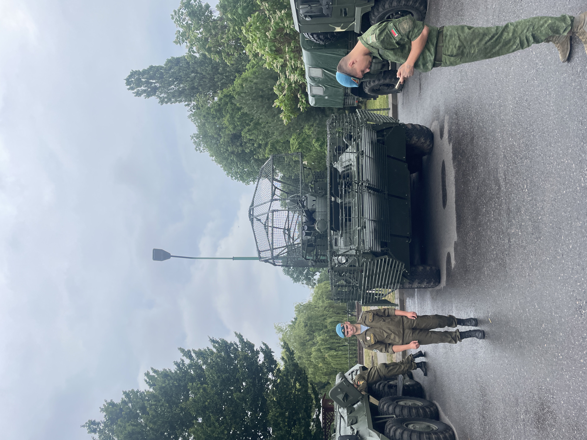 In an outdoor, overcast setting, likely in Brest, Belarus, two individuals in military uniforms stand near armored vehicles. The person on the left, wearing a light brown uniform and a blue beret, stands facing forward with a neutral expression. To the right, another soldier in a camouflage uniform, also wearing a blue beret, is looking down at their mobile phone. Behind them are at least two large, green military vehicles, one appearing to be a radar system with a prominent dish, and another resembling a truck. The ground is wet, suggesting recent rain. Trees and foliage are visible in the background, contributing to the park-like or outdoor military exhibition atmosphere.