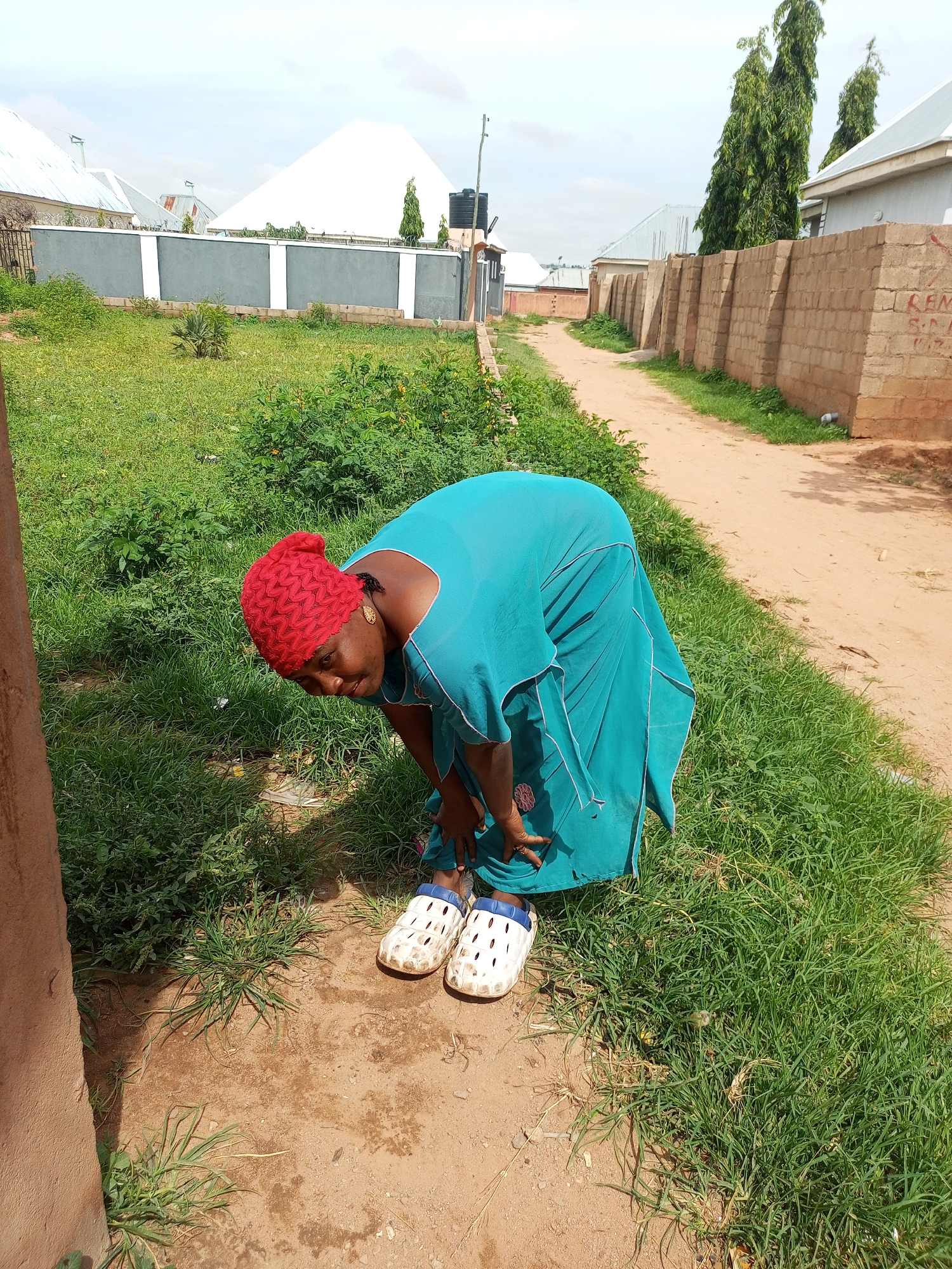 In Tempure, Nigeria, a woman wearing a bright red headscarf and a teal-colored dress is bending over in a grassy area next to a dirt path. She is wearing white Crocs-style shoes. The woman appears to be reaching for something on the ground. In the background, there are several buildings and a brick wall along the side of the path. The sky is overcast.