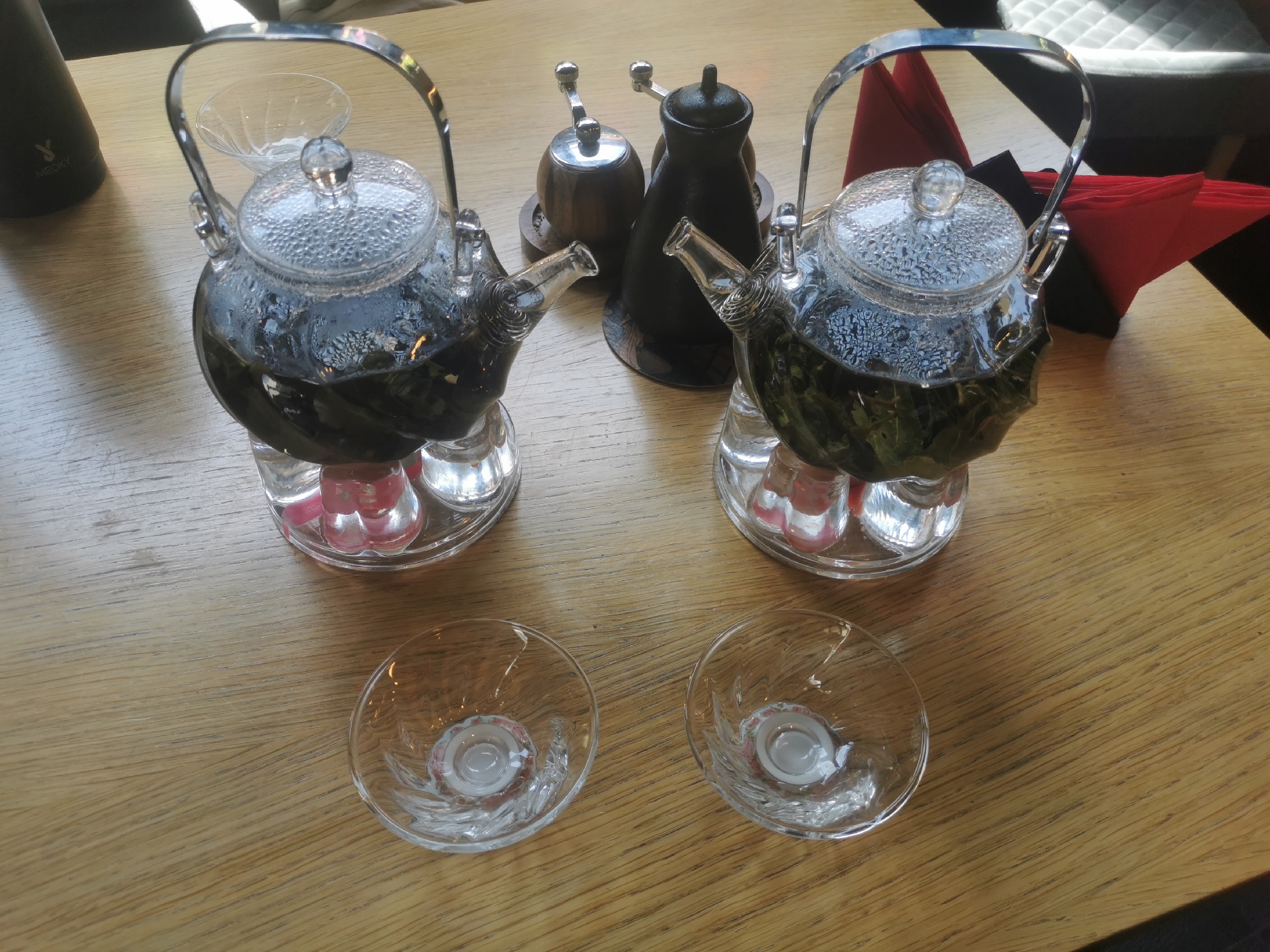 An eye-level shot captures a dining table set for two, featuring a pair of identical glass teapots filled with brewing tea, likely in a cafe or restaurant in Blagoevgrad, Bulgaria. The teapots, made of clear glass with metal handles and textured glass lids, show visible condensation, indicating the tea is hot. Green tea leaves are clearly visible steeping inside each teapot. Both teapots rest on individual clear glass bases, which appear to house small, red-glowing warmers, possibly tealights, to maintain the tea's temperature.
In front of the teapots are two small, ornate clear glass bowls or cups, ready for serving the tea. In the background, between the two teapots, a pair of condiment shakers are visible: one with a wooden body and metal top, and the other a darker, possibly ceramic or metal, bottle-shaped shaker. To the far left, a dark, conical thermos or bottle with a white "MIROKY" logo and a stylized "Y" is partially visible. On the right side, folded red and black napkins are neatly stacked, with a glimpse of a gray padded chair in the blurred background. The table surface is light-colored wood with a distinct grain pattern. The overall scene suggests a calm and inviting tea-drinking experience.