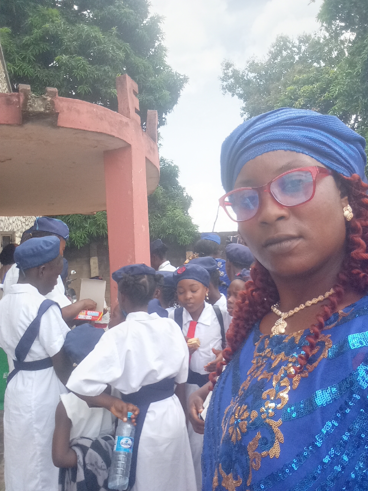 A photograph taken in Akwanga, Nigeria, depicts a woman in the foreground wearing a blue, sequined top and matching headwrap, as well as red-framed glasses and a gold necklace and earrings. Her hair is styled in auburn-colored spiral curls. In the midground, a group of children wearing matching white uniforms with dark blue sashes and caps are gathered. Some are holding water bottles and snacks. The setting appears to be outdoors, near a building with a red column and overhanging structure. Trees are visible in the background.