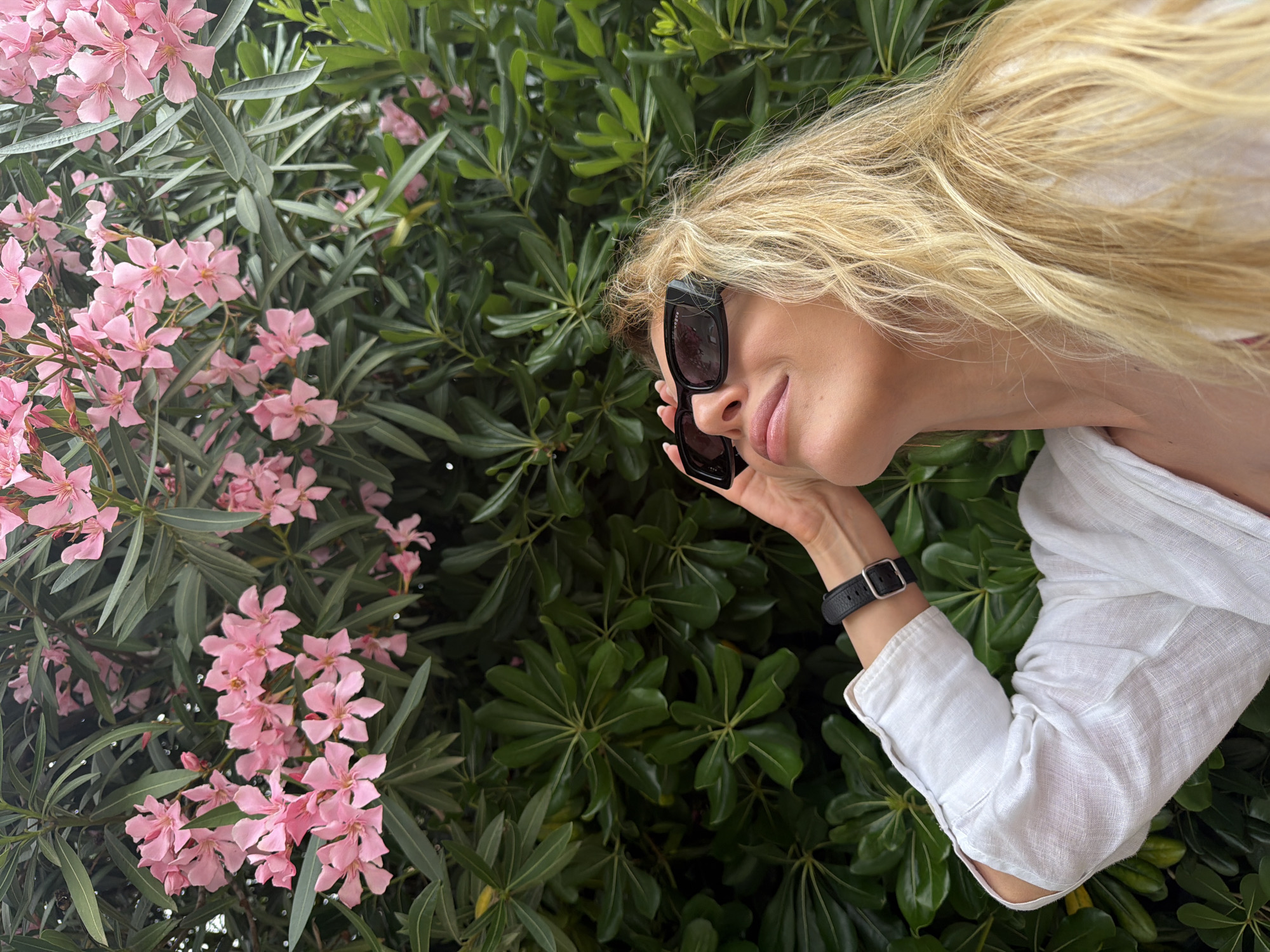 A woman with long, flowing blonde hair is partially visible, peeking out from behind dense green foliage adorned with clusters of pink flowers. The image is captured in a diagonal orientation, with the woman's head tilted to her right, looking downwards with a subtle smile. She wears large, black-framed rectangular sunglasses, which she lightly touches with her right hand. Faint text such as "SUNGLASSES" and "UV" is discernible on the inside arm of the sunglasses. A black-strapped watch with a silver buckle is visible on her left wrist. She is dressed in a white, textured long-sleeved shirt, possibly linen.
The background is a vibrant tapestry of dark green leaves and numerous five-petaled pink flowers, consistent with plants like Oleander, commonly found in Mediterranean regions. The scene is outdoors, well-lit by natural daylight, suggesting fair weather. The overall mood is relaxed and casual, portraying an individual enjoying a natural, floral environment in Budva, Montenegro.