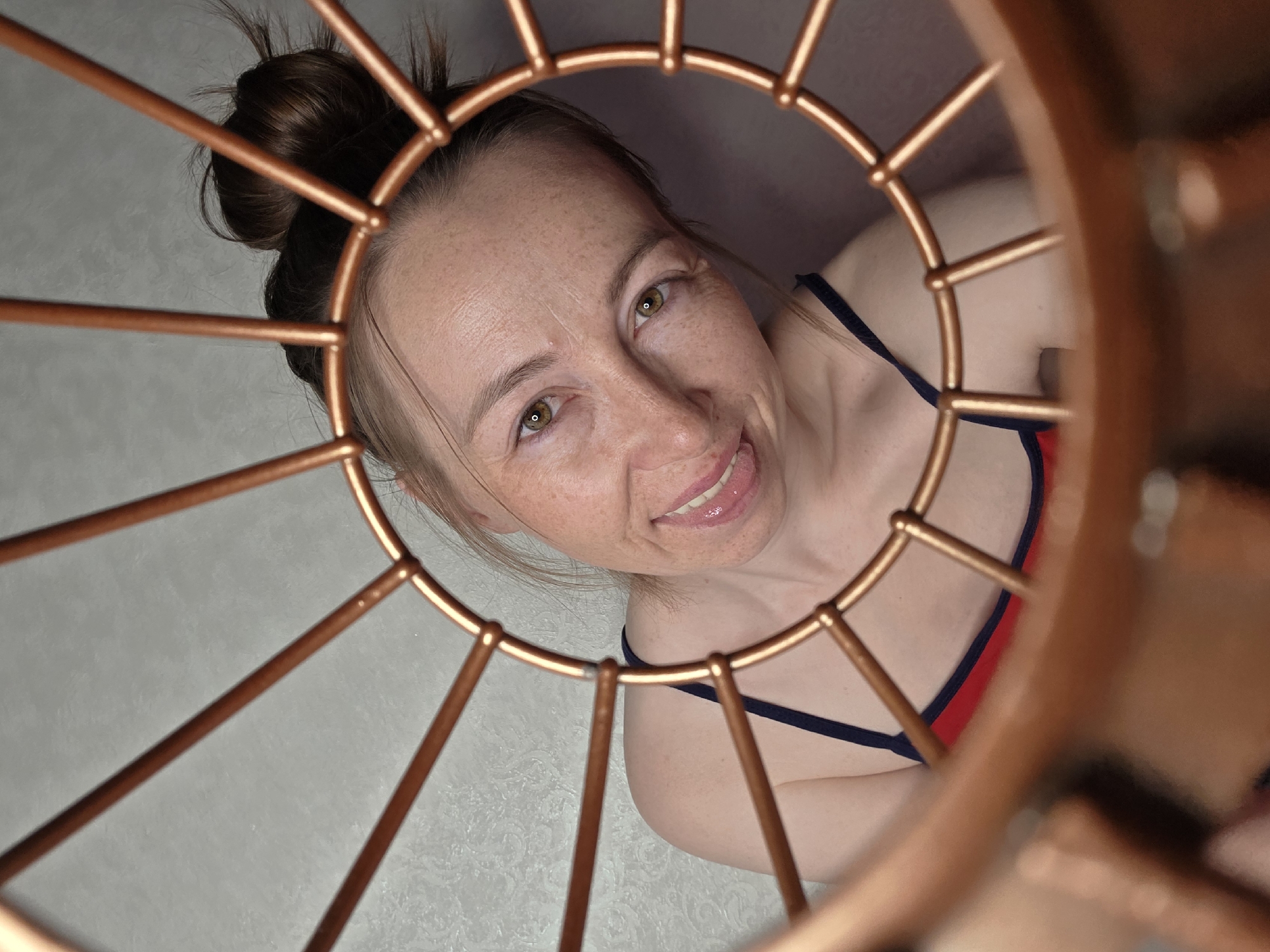A woman is seen from a low-angle perspective, looking up through a copper-colored, circular structure with radiating spokes. She has fair skin, freckles, and light brown hair pulled up in a messy bun. She is wearing a red top with navy blue straps and is smiling slightly, sticking out her tongue. The background appears to be a light gray or white textured surface. No specific location is discernible from the image to determine if it was taken in Chaikovsky, Russia.