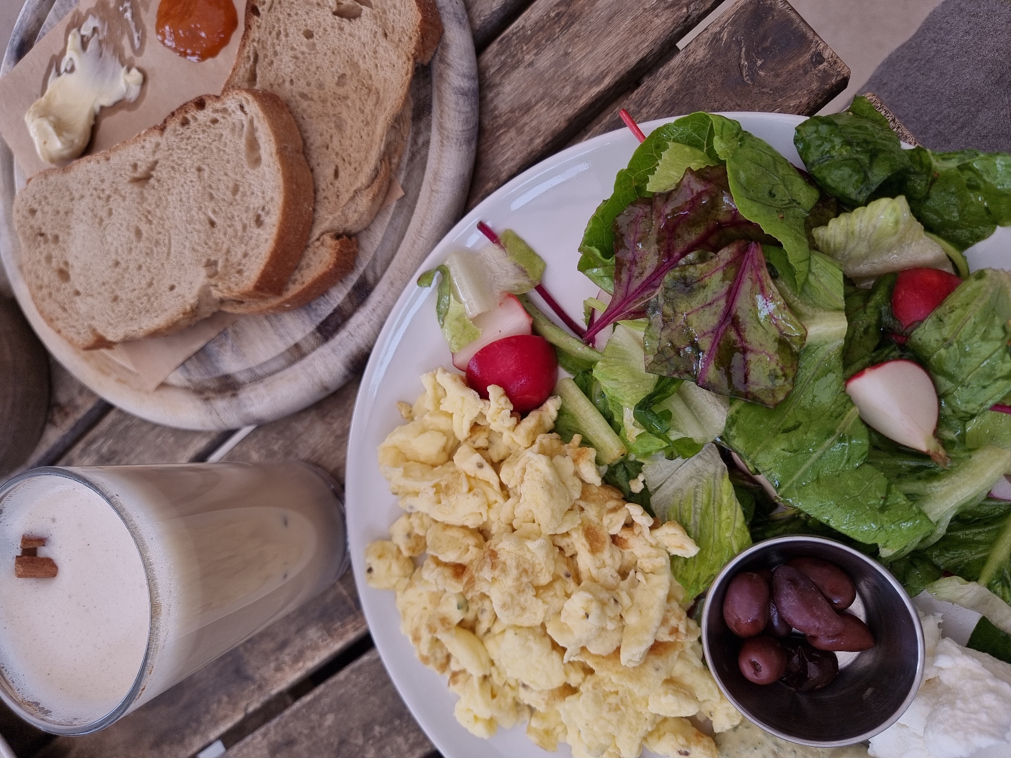 A table at a restaurant in Mitzpe Ramon, Israel is set with a breakfast spread.
A wooden plate holds three slices of wheat bread, accompanied by butter and jam served on a piece of parchment paper.
A separate white plate presents a meal comprising scrambled eggs, a mixed salad with radishes, olives in a small metal bowl, and a dollop of white cheese.
A tall glass contains a milky beverage topped with a cinnamon stick, sits near the plate.