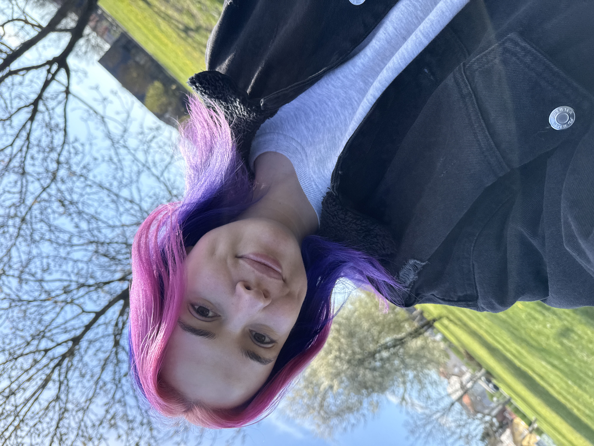 A low-angle, eye-level shot shows a person with pink and purple hair, fair skin, and visible eyebrows looking towards the camera. They are wearing a light gray top and a black jacket with a shearling-lined collar. The background is a blurred outdoor setting with green grass, trees, and a blue sky. Bare tree branches are visible against the sky. The image appears to have been taken in a park-like setting in Zagreb, Croatia, as evidenced by the distinctive buildings and landscape typically found there. No specific street names or locations can be determined from this image.