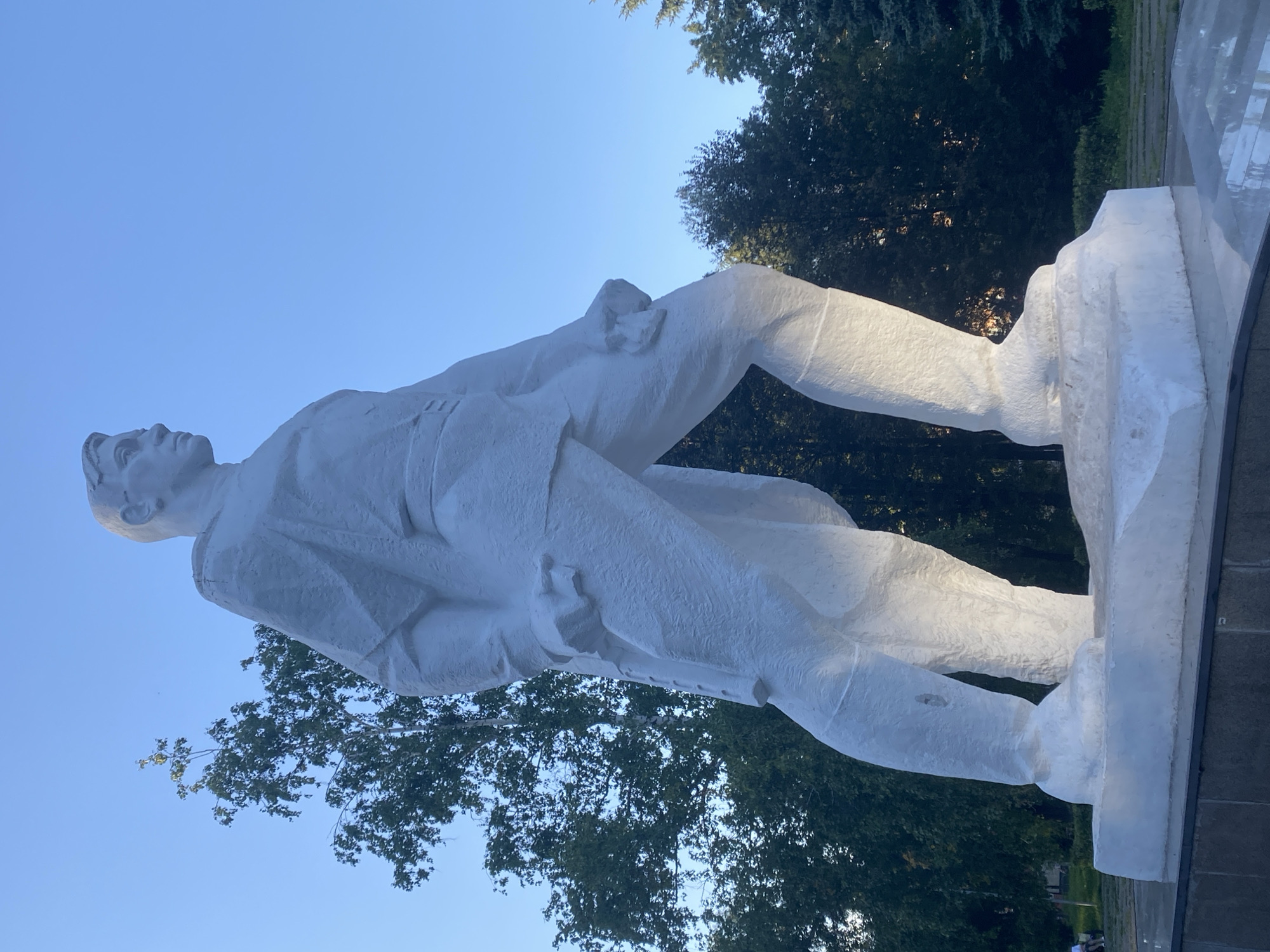The image shows a white, full-body statue of a man in military uniform, standing on a pedestal. The statue is made of a rough, textured material. The man is facing to the right in profile, with his head turned slightly towards the viewer. He is wearing a tunic with a belt and trousers tucked into tall boots. His left leg is forward, and his arms are held down at his sides, with his hands appearing to hold something. The background consists of a clear blue sky, several trees, and what appears to be a park-like setting. The statue is located in Novokuznetsk, Russia. Specifically, it is a monument to the Soviet pilot Viktor Vasilyevich Talanov, located on Talanova Street in the Zheleznodorozhny District of Novokuznetsk. The monument is part of a memorial complex.