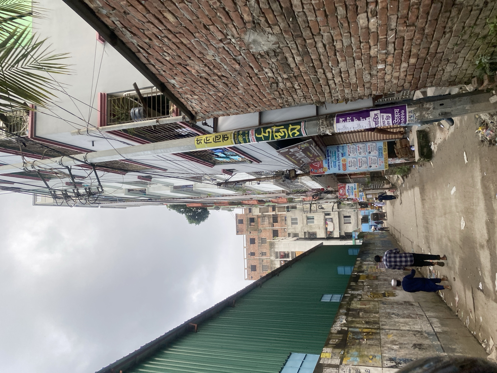 This is a vertical, eye-level shot looking down a narrow alleyway in Dhaka, Bangladesh, flanked by tall, multi-story buildings. On the left is a long, green corrugated metal structure with a sloped roof. The wall to the left of the alley is covered in faded posters. In the center of the alley, two males are walking away from the camera. The one in front is wearing a blue thobe and a white kufi, and the one behind him is wearing a plaid shirt and dark pants. Further down the alley, other people and a rickshaw are visible. The building on the right is made of brick and has multiple windows, some with metal bars. A utility pole with various signs and wires is attached to the building. The sky is overcast.