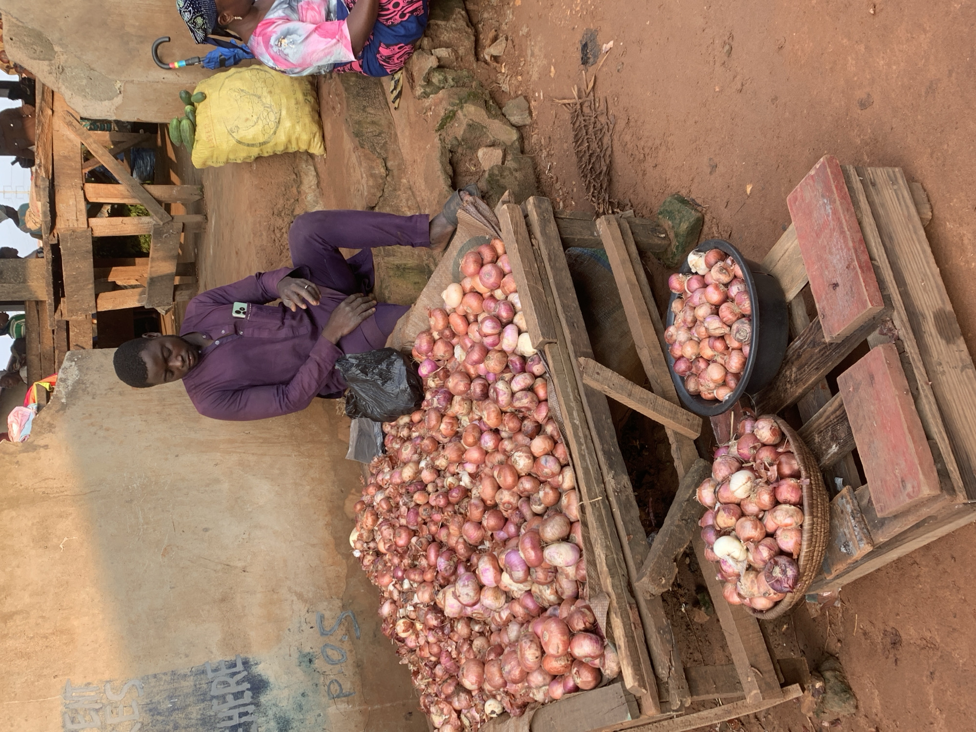 A young Black man in a purple tunic and pants sits on a step beside a large pile of red onions. He is looking down. To his right, a Black woman in a colorful patterned top and headwrap is also seated. Behind the man is a rough, textured wall with some faint blue and white markings. A yellow sack filled with what appear to be cucumbers sits near the woman. In front of the man are two baskets and a black bowl, all filled with onions, placed on wooden crates. The ground is reddish-brown dirt. The scene appears to be a market stall in Shagamu, Nigeria.
