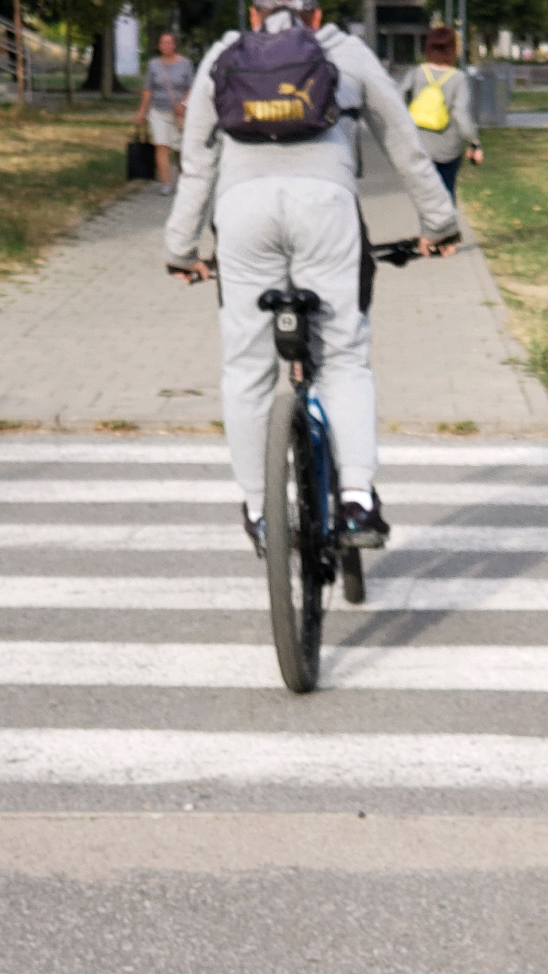 A person is riding a bicycle across a pedestrian crossing. The person is wearing a gray hooded sweatshirt, light gray pants, and a dark backpack with a Puma logo. They are also wearing dark shoes with white socks. The bicycle has a dark frame and is viewed from behind, with the rear wheel prominently in the foreground. The pedestrian crossing has white stripes on an asphalt road. In the background, two women are walking on a paved sidewalk. One woman on the left is wearing a gray top and a white skirt and carrying a black bag. The woman on the right is wearing a gray top and blue pants and a yellow backpack. The environment appears to be an outdoor urban setting with trees and buildings visible in the distance. The image quality is slightly blurred. This scene could be located in the city of Prešov, Slovakia.