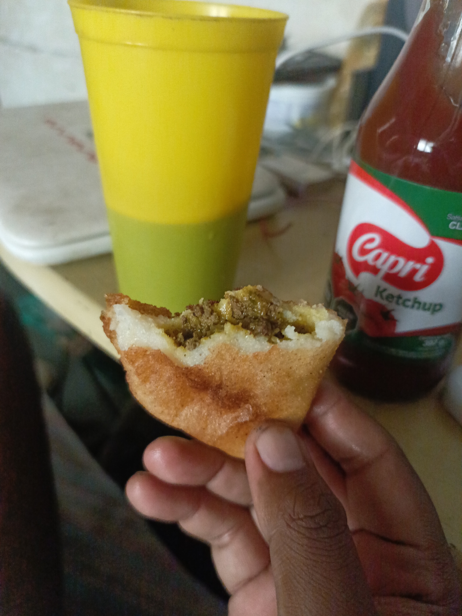 A partially eaten arepa filled with seasoned ground meat is being held by a dark-skinned hand. A tall yellow plastic cup is in the background to the left. A bottle of Capri brand ketchup is to the right. The scene appears to be indoors, likely at a table. The location is Juan Griego, Venezuela, as indicated by the Capri Ketchup brand, which is commonly found in Venezuela.