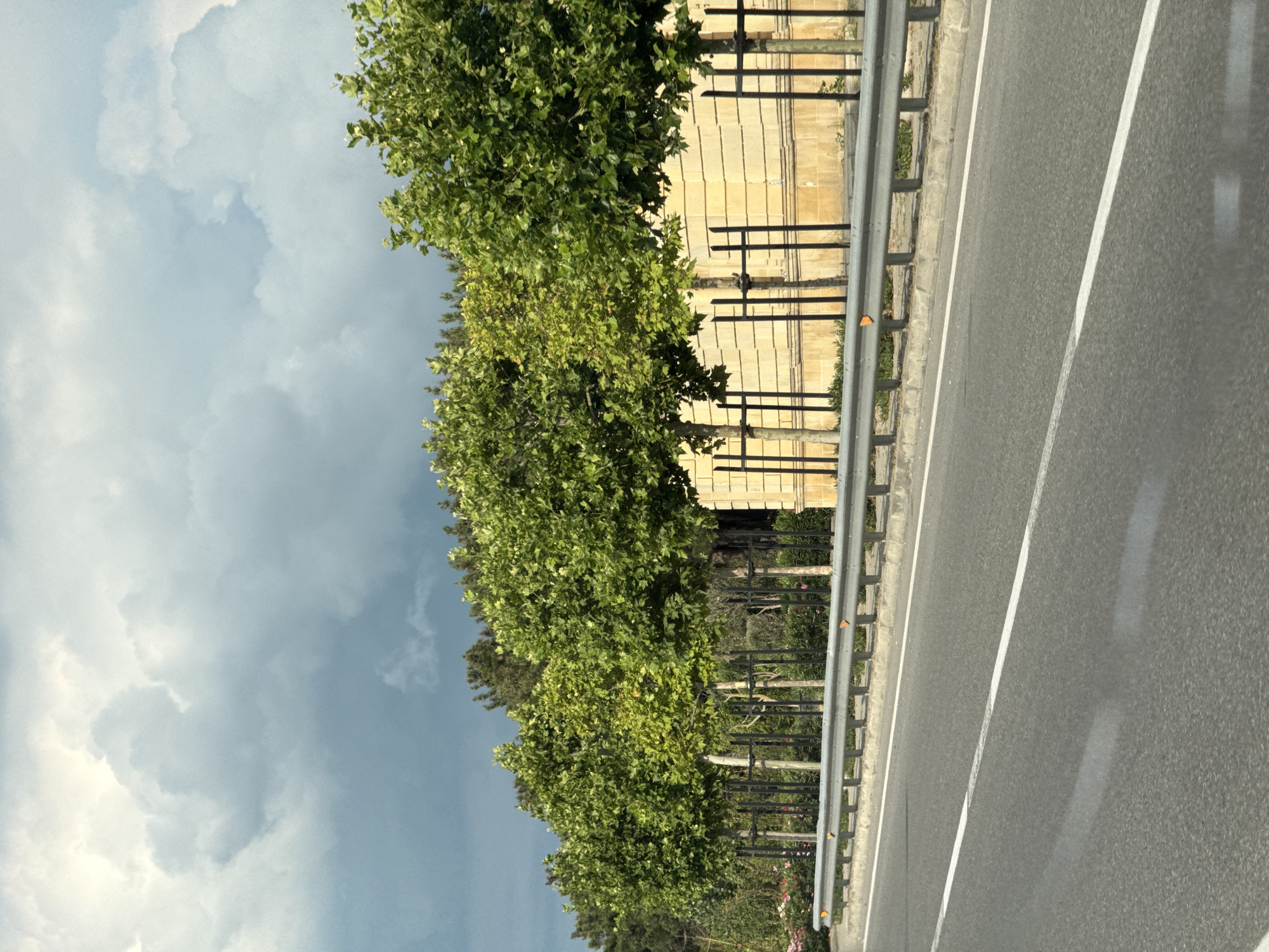 The image captures a roadside scene on a slightly overcast day, likely in the Khazar Raion region of Azerbaijan. The foreground is dominated by an asphalt road with clear white lane markings, indicating a highway or major road. A silver metal guardrail runs along the side of the road, separating it from a paved shoulder and a strip of greenery.
Beyond the guardrail, a row of lush green trees with dense foliage lines the edge of the road. Behind the trees, a light-colored building with vertical lines, possibly a wall or facade, is visible. The building appears to be constructed from large, rectangular blocks. The sky is filled with dramatic clouds, suggesting a change in weather, with pockets of bright light peeking through. The overall impression is that of a scenic drive through a developed area with natural elements. There is no visible text in the image.