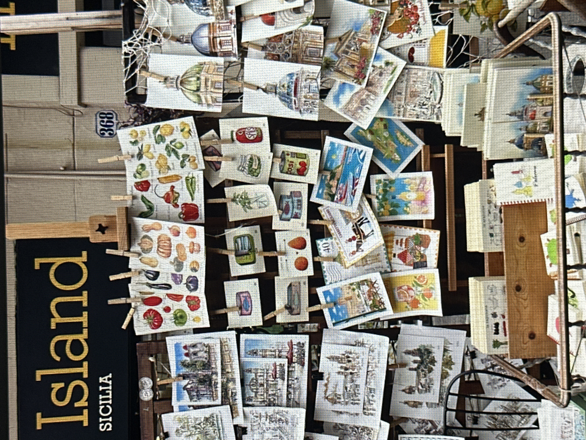 The image displays a close-up, overhead view of a wall adorned with numerous small, framed pictures, possibly postcards or prints. These are attached to the wall by clothespins, creating a layered and somewhat chaotic display. The pictures themselves feature a variety of subjects. Many depict architectural scenes, including domes, intricate buildings, and cityscapes, some with a classical or European aesthetic. Other pictures showcase still-life arrangements, such as colorful illustrations of fruits and vegetables, jars of preserves, and what appear to be decorative cans. There are also images of landscapes with water and sky, and one notable postcard featuring a sailboat.
A black banner with large, gold, sans-serif lettering is visible on the left side of the image. The word "Island" is written vertically in large, bold letters, followed by the word "SICILIA" in smaller, capitalized letters below it. This strongly suggests a Sicilian theme for the displayed items.
Other details include a small blue oval tag with the number "368" attached near the top left. The lighting suggests it could be daytime, though the specific time of day is unclear. The overall impression is of a souvenir shop or a display of local art and crafts, possibly in a market or along a street in Sicily, given the prominent "SICILIA" text. The arrangement of the pictures, attached with clothespins, gives a casual and artisanal feel to the presentation. There are no people visible in the image.