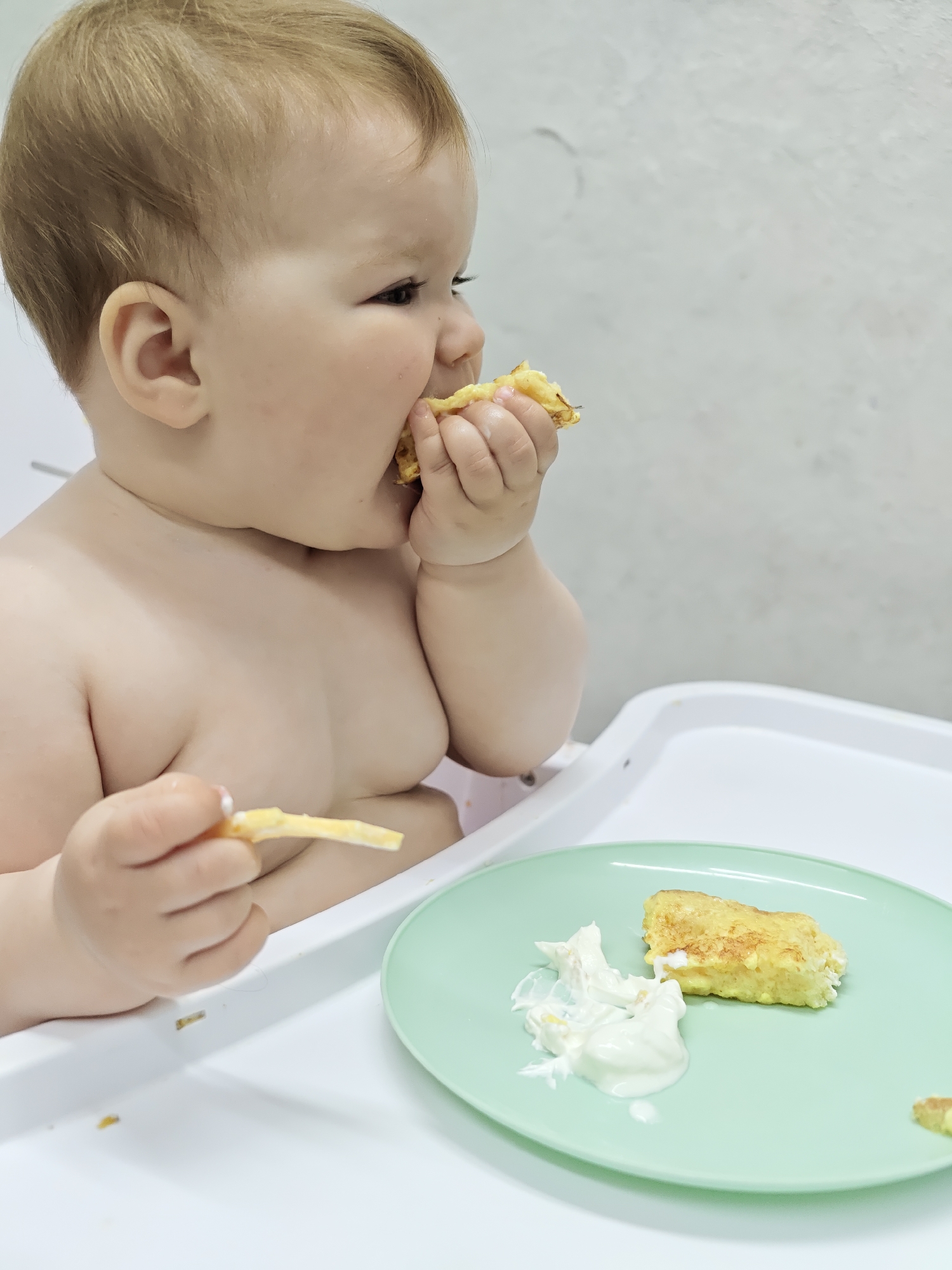 The image depicts a fair-skinned infant with light brown hair seated in a high chair. The baby is eating a piece of what appears to be fried food or an omelet. A green plate on the high chair tray contains a portion of the same food and a dollop of white sauce. The background is a plain, light-colored wall. The location is reported as Chaikovsky, Russia.