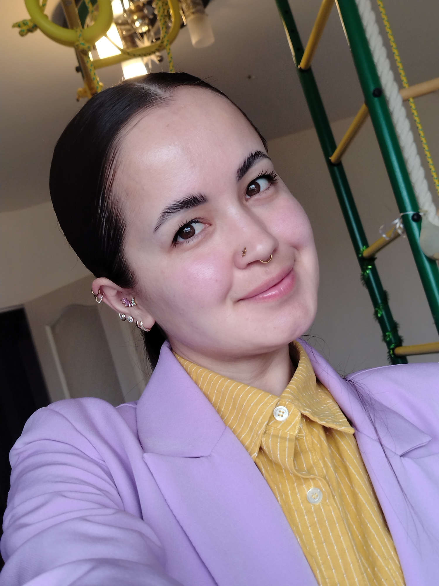A woman is posing for a selfie in an interior setting. She has dark hair pulled back, light skin, and multiple piercings in her left ear and nose. She is wearing a yellow striped collared shirt and a lavender blazer. Behind her is a green and yellow play structure with a rope ladder. The background suggests an indoor space, possibly a home. The location in the city of Noviy, Russia is unknown.