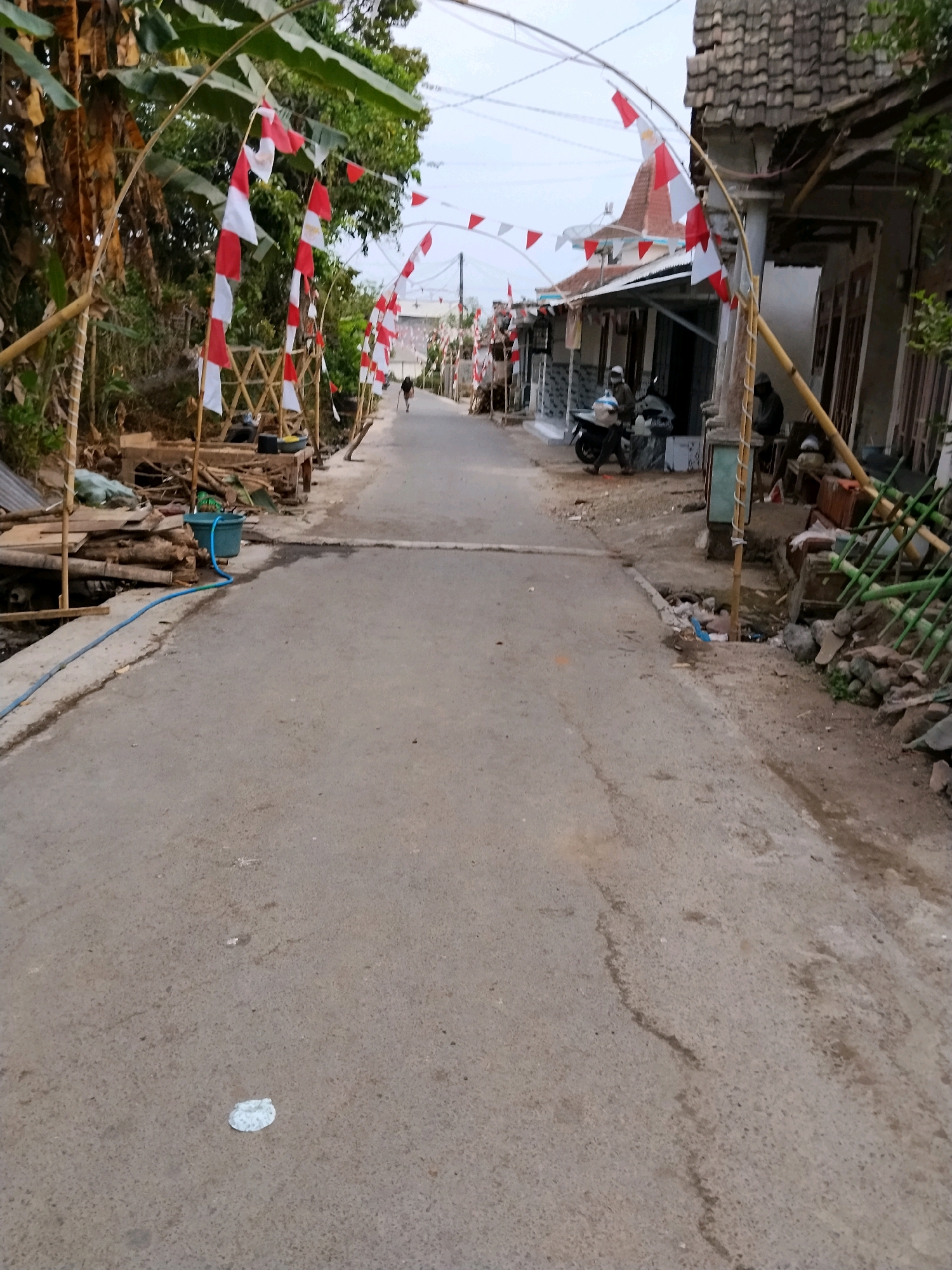 The image depicts a narrow, paved street in Tlogosari, Indonesia, lined with residential structures and foliage. The street is decorated with alternating red and white triangular flags, strung along bamboo arches that span the road. On the left side of the street, a pile of lumber and building materials is visible, alongside a blue plastic container with a blue hose connected to it. A single person is walking away from the camera down the street. On the right side of the street, a motorcycle is parked in front of a house. Two individuals are seated near the front of the house. The sky is overcast.