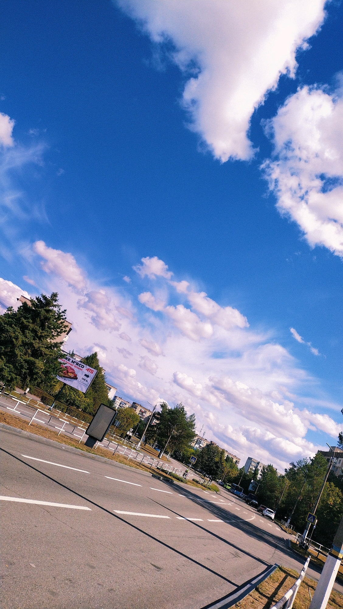 A tilted, full-shot outdoor photograph captures a bright, sunny day in Horishni Plavni, Ukraine. The scene looks down upon a paved road with white lane markings, leading into the distance. On the right side of the road, a yellow and blue pole stands near the curb, with a pedestrian crossing sign visible behind it. Further down the road, a few cars are parked and driving. To the left of the road, a billboard displaying meat products and the ATB supermarket logo is visible. Behind the billboard and road, there are trees and buildings. The sky above is a vibrant blue, dotted with fluffy white clouds. The overall impression is of a typical urban landscape bathed in sunlight.