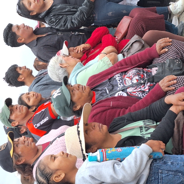 A group of people of varying ages are looking upwards. Most are wearing casual clothing, including hats and sweaters. A young girl in the foreground holds a brightly colored tube. The crowd appears to be densely packed together, possibly observing something in the sky. Their expressions suggest anticipation or curiosity.