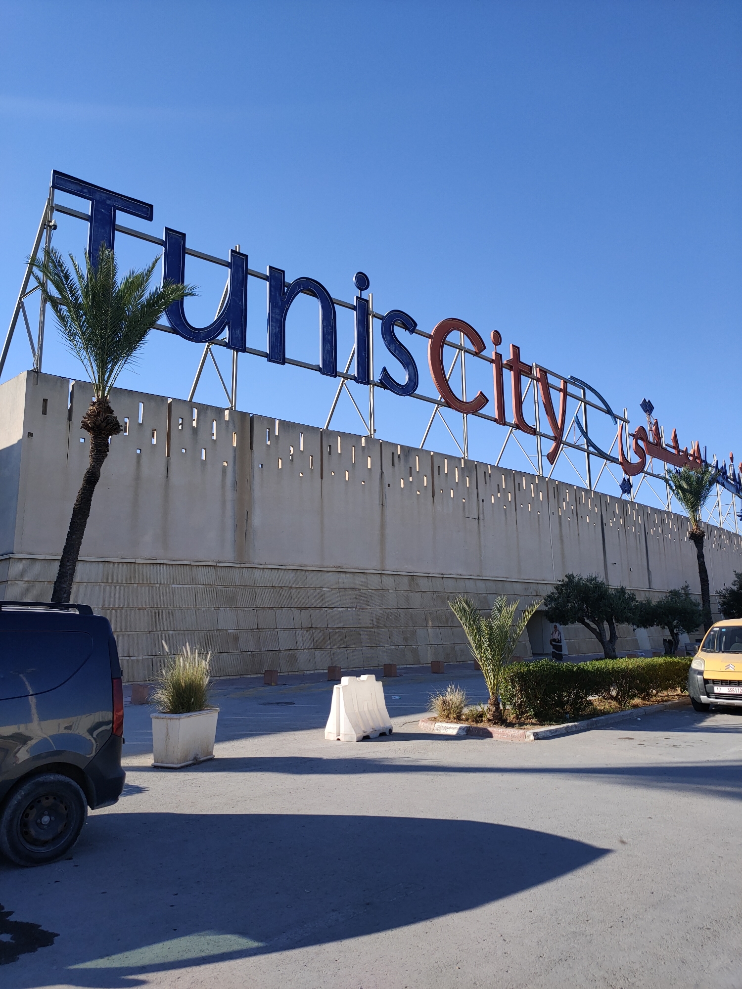 The image shows a large sign that reads "TunisCity" in blue and red letters, with Arabic script also visible to the right. The sign is situated in front of a tan-colored building with a textured facade. To the left of the sign, a palm tree leans towards the right. In the foreground, a dark-colored van is parked on the left side of the frame, and a yellow car is parked on the right. A white plastic barrier is visible near the yellow car. The ground is paved, and shadows are cast across it, indicating a sunny day. The sky is clear and blue. The setting appears to be an outdoor commercial or entertainment complex, likely located in Cebalat, Tunisia, given the "TunisCity" signage which indicates a prominent location in the capital city region.