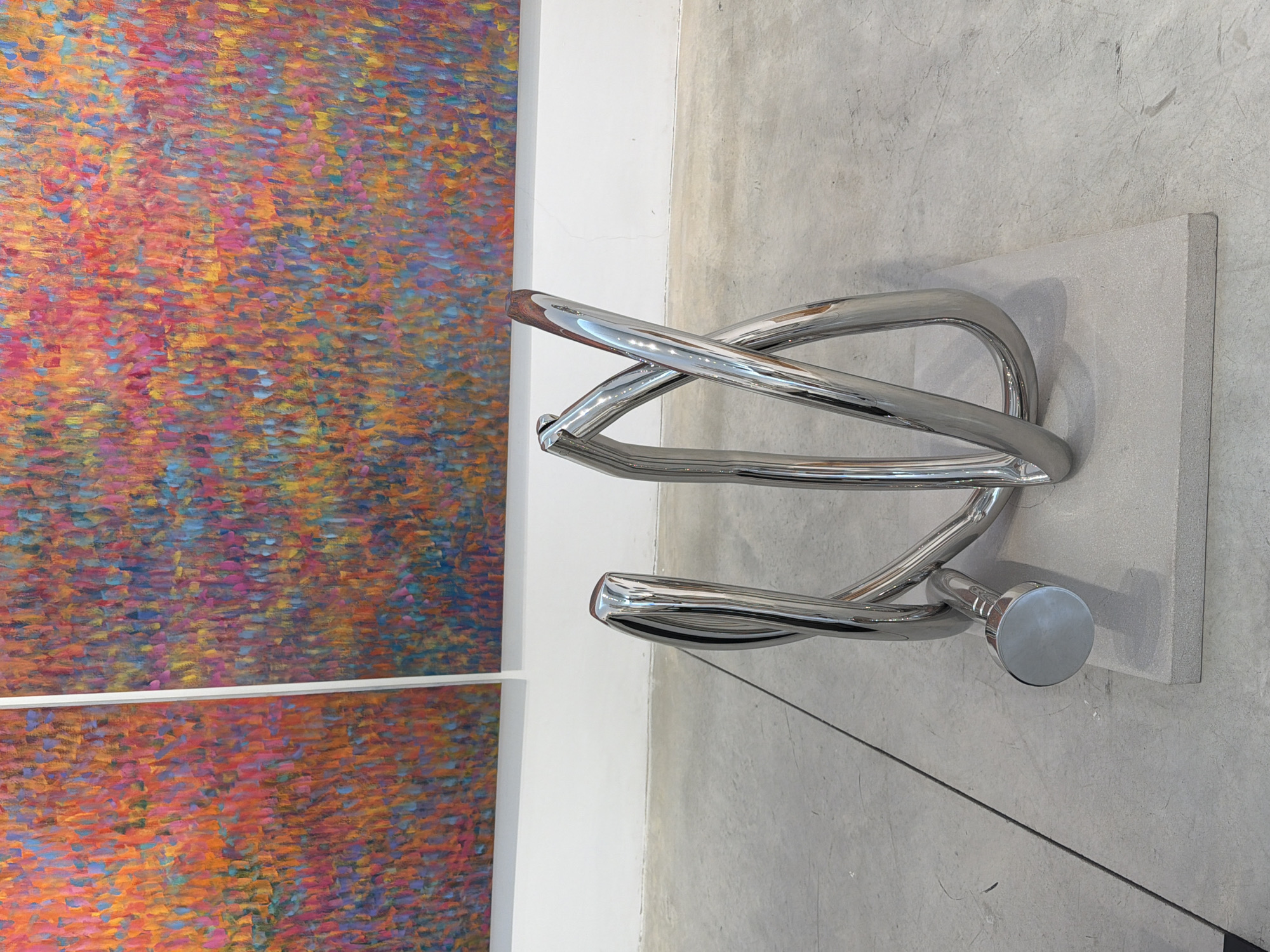 The image depicts a contemporary art exhibition at the Tel Aviv Museum of Art. In the foreground, a polished chrome sculpture is displayed on a light gray rectangular plinth, which rests on a concrete floor. The sculpture's form is abstract, with tubular elements twisted and intertwined, resembling a metallic knot or a stylized hairpin. The chrome surface reflects the surrounding environment. In the background, two large, vibrant abstract paintings hang on a white wall. The paintings feature a textured surface composed of numerous small brushstrokes in a palette of reds, oranges, pinks, blues, and yellows, creating a sense of energetic movement. The overall impression is one of modern artistic display, with a focus on the interplay of form, color, and light.