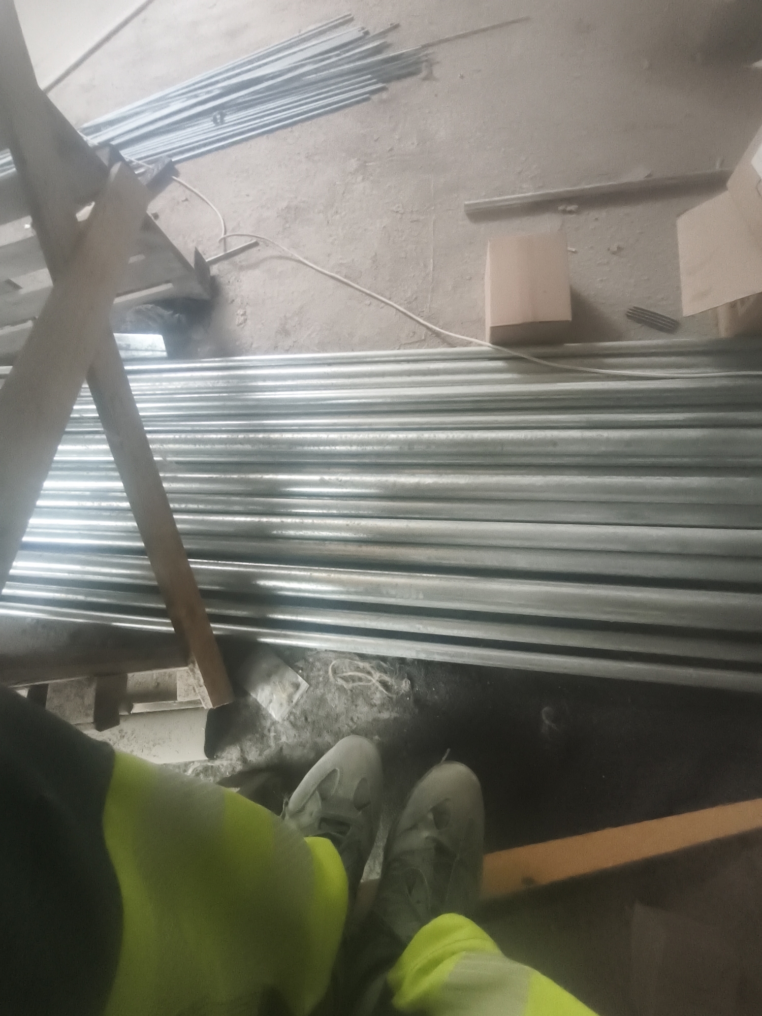 The image depicts a person wearing high-visibility work attire, including a neon yellow and black jacket and grey work boots, standing in what appears to be a workshop or construction area. The foreground is dominated by the person's legs and feet, positioned over a wooden beam. In the background, a large stack of metal pipes is visible, arranged horizontally. The environment is industrial, with a concrete floor and evidence of construction materials such as cardboard boxes and wooden supports. A light-colored electrical cord snakes across the floor near the pipes. The overall scene suggests a work-in-progress environment, possibly within a warehouse, manufacturing facility, or construction site. No specific landmarks or context point to the city of Riga, Latvia.