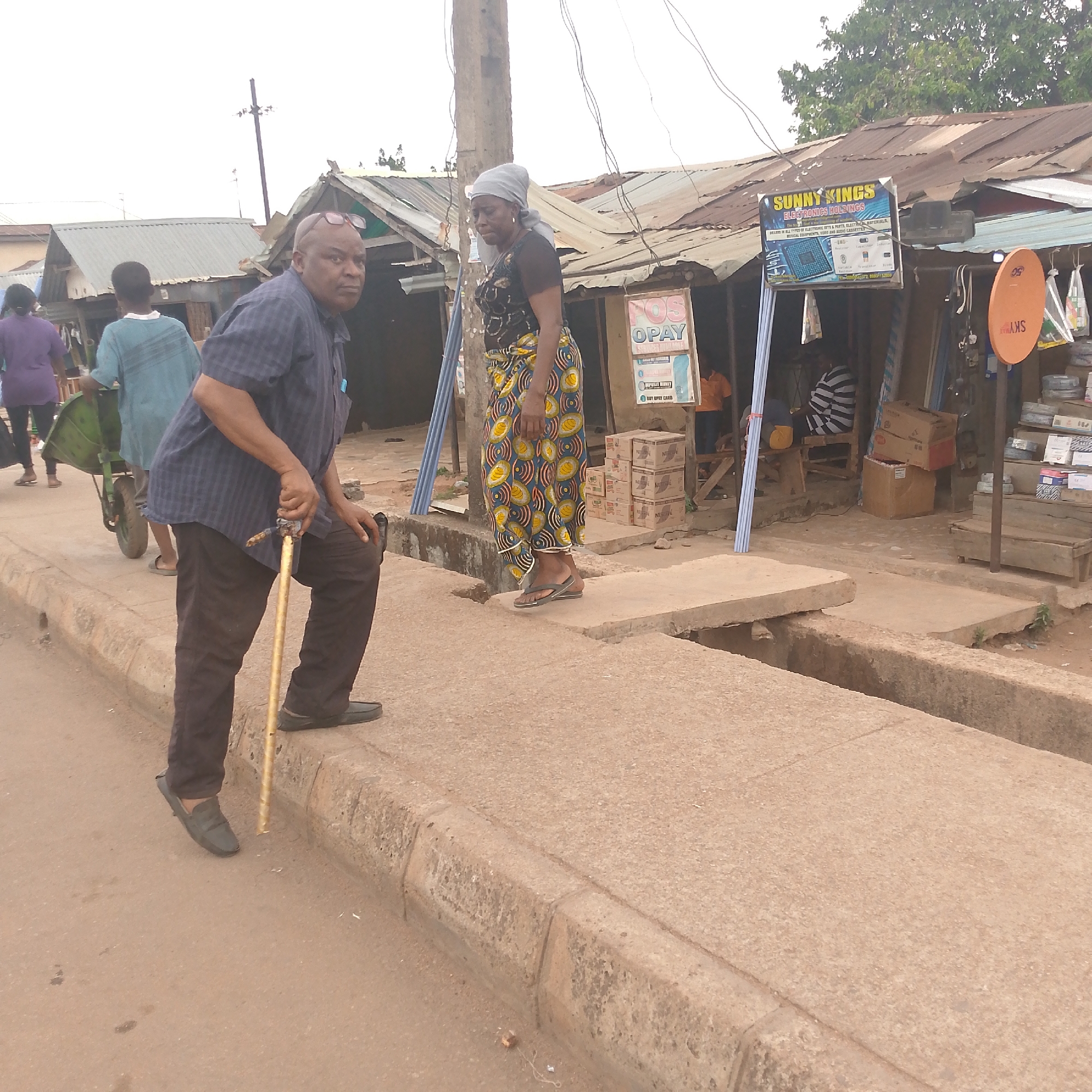 The image shows a street scene in Bukuru, Nigeria. An elderly man with glasses and a blue checkered shirt stands on the sidewalk with a golden walking stick. A woman wearing a headscarf, a black t-shirt, and a patterned yellow and blue skirt is standing on a concrete platform that appears to cross a ditch. In the background, there is a small shop with a corrugated metal roof. The shop has a sign that reads "SUNNY KINGS ELECTRONICS HOLDINGS." Another sign reads "OPAY." Several people are visible in the shop and on the street. There is a wheelbarrow beside a man with a green shirt, on the left side of the image.