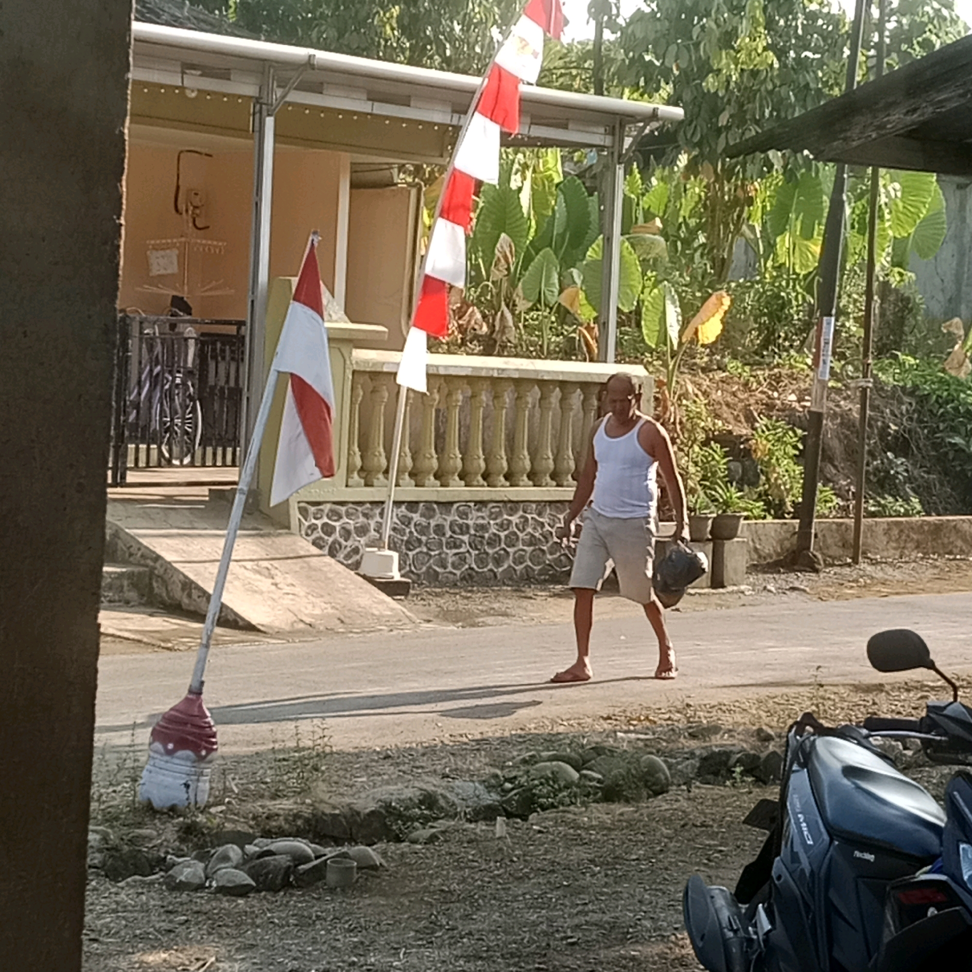 In Rakit, Indonesia, a man wearing a white tank top and light-colored shorts walks on a paved road. He is carrying a dark bag in his right hand. A red and white Indonesian flag on a pole is planted to his left. The flag is made of alternating red and white rectangular sections. In the background, there is a building with a covered porch and lush green foliage. A motorcycle is parked in the foreground to the right. The ground appears to be a mix of dirt and gravel.