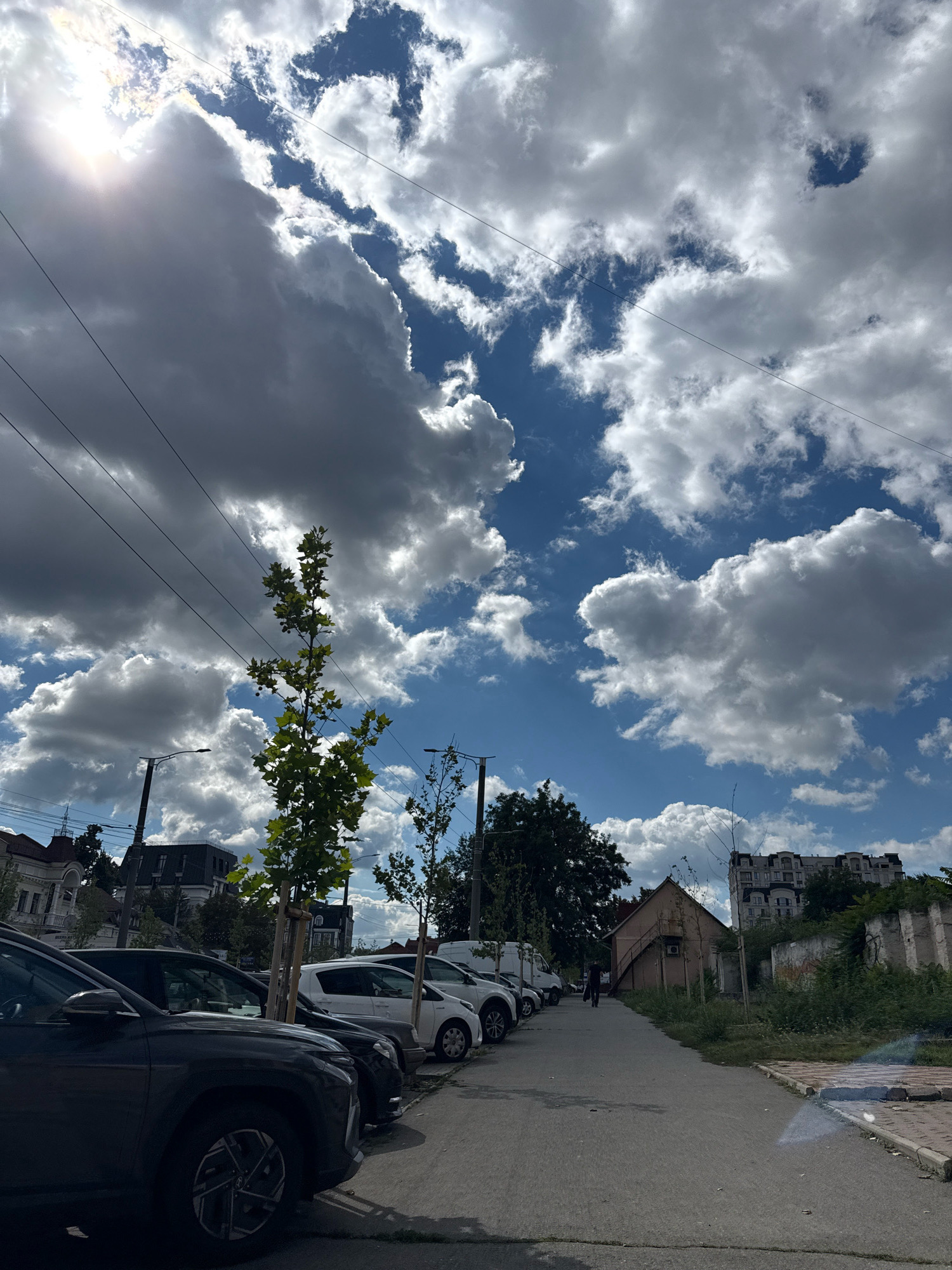The image depicts a street scene in Chișinău, Moldova, under a dynamic sky with a mix of dark and bright clouds, and the sun peeking through. A row of parked cars is visible on the left side of the paved road, including a dark-colored SUV in the foreground. Beyond the cars, there are buildings of various architectural styles, some appearing older and residential, while others are more modern. Trees are planted along the street, and utility lines are strung overhead. A lone individual is walking away from the viewer on the right side of the road, which is bordered by overgrown vegetation and a crumbling wall. The overall impression is that of a somewhat neglected urban environment on a partly cloudy day.