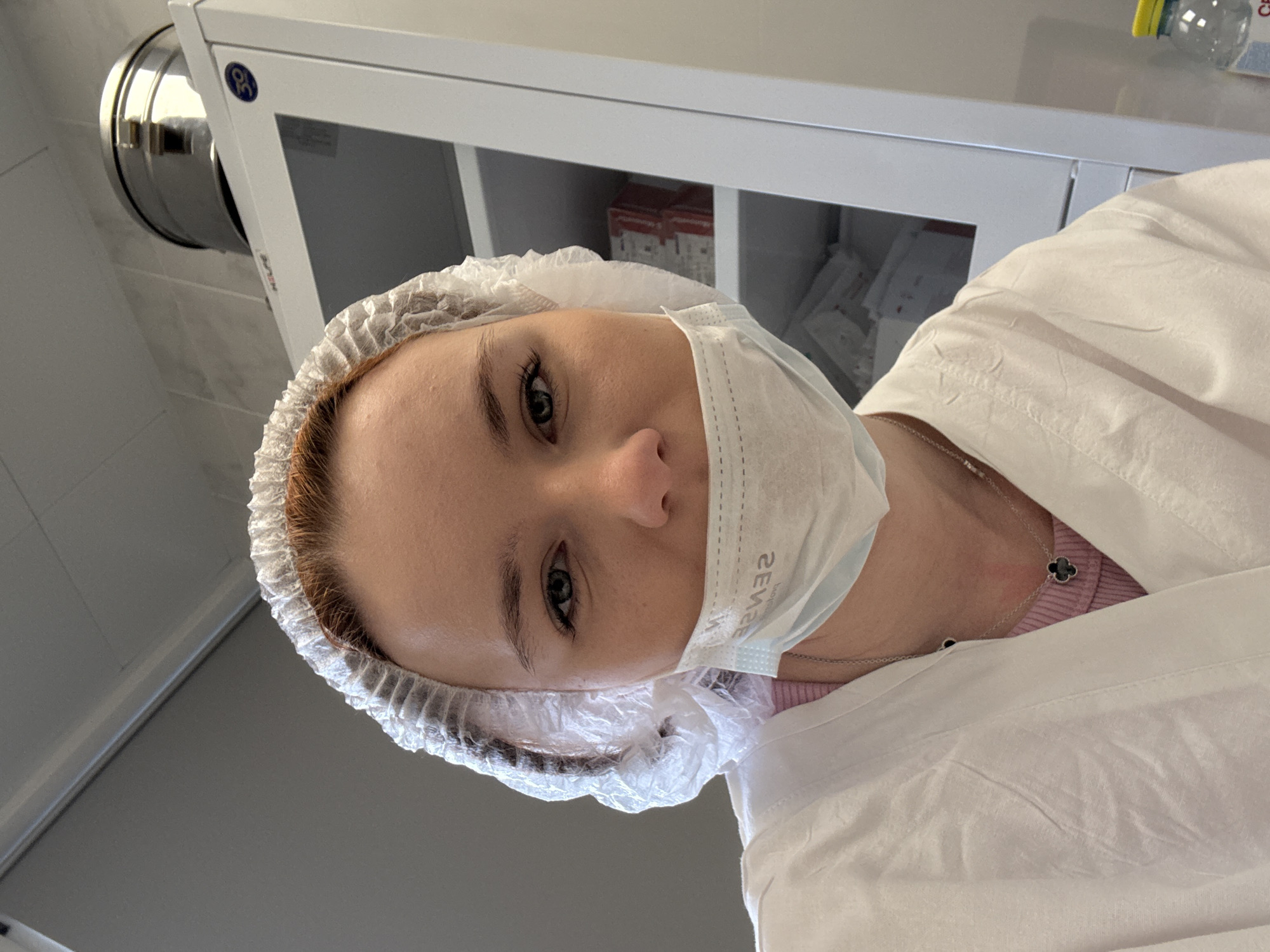 A person with fair skin, wearing a white bouffant cap and a light blue disposable face mask, looks directly at the camera. The mask partially obscures the lower half of their face. They are also wearing a white lab coat over a pink top. A silver necklace with a four-leaf clover pendant is visible around their neck.
The background includes a white cabinet with items stored inside. A rounded metal object is attached to the wall behind the cabinet. The location is unidentified. The city and country are also unidentified.