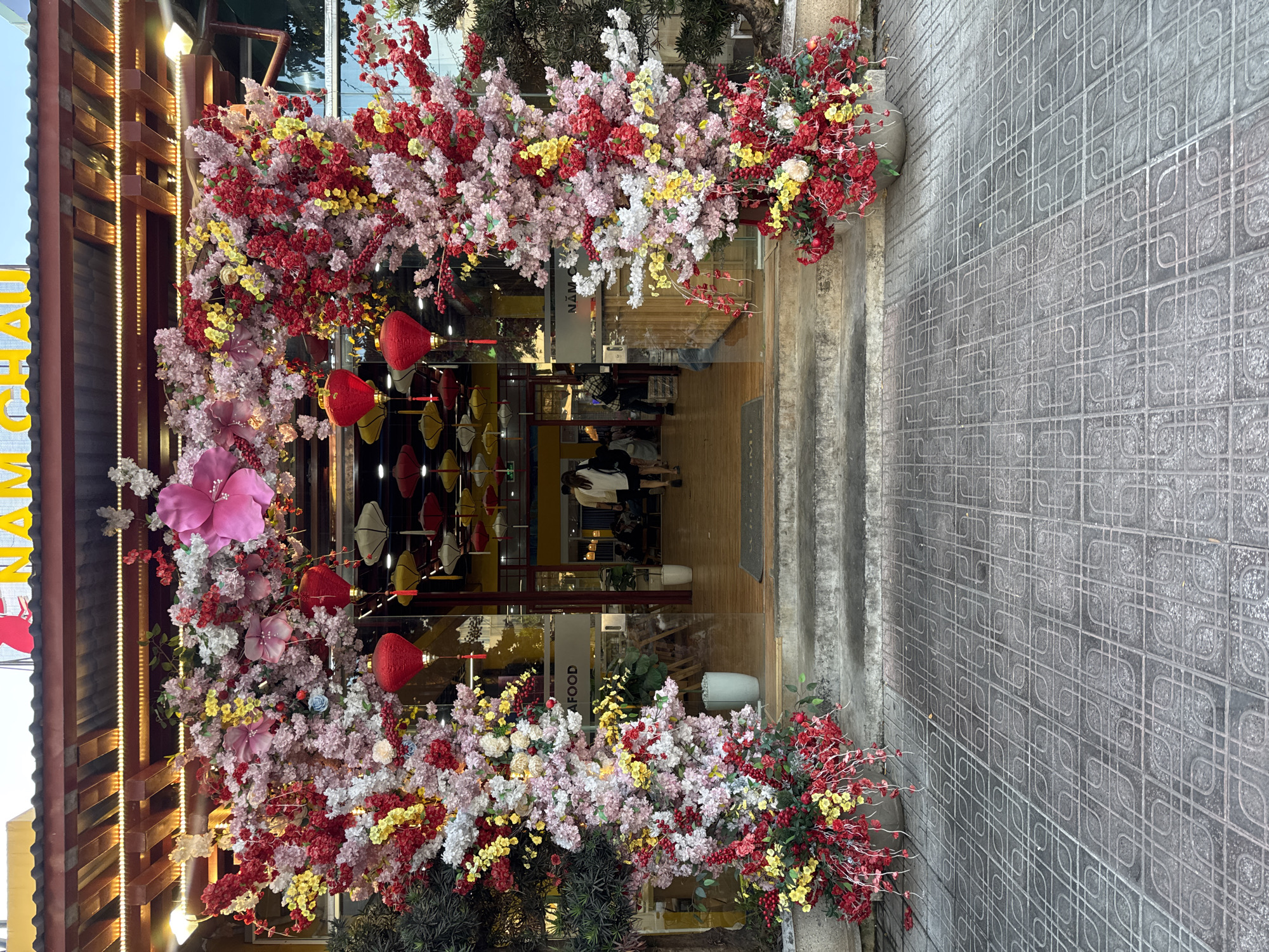 An exterior shot depicts the entrance to a Vietnamese restaurant. The entrance is elaborately decorated with an arch of flowers in shades of red, yellow, pink, and white. Red lanterns are hung along the archway. The restaurant's name, "NAM CHAI," is visible in large yellow letters on the building's facade. Through the glass entrance, several people are seated at tables inside the restaurant. The exterior sidewalk consists of gray patterned tiles. The location is likely within Đà Nẵng, Vietnam, given the restaurant's name and style.