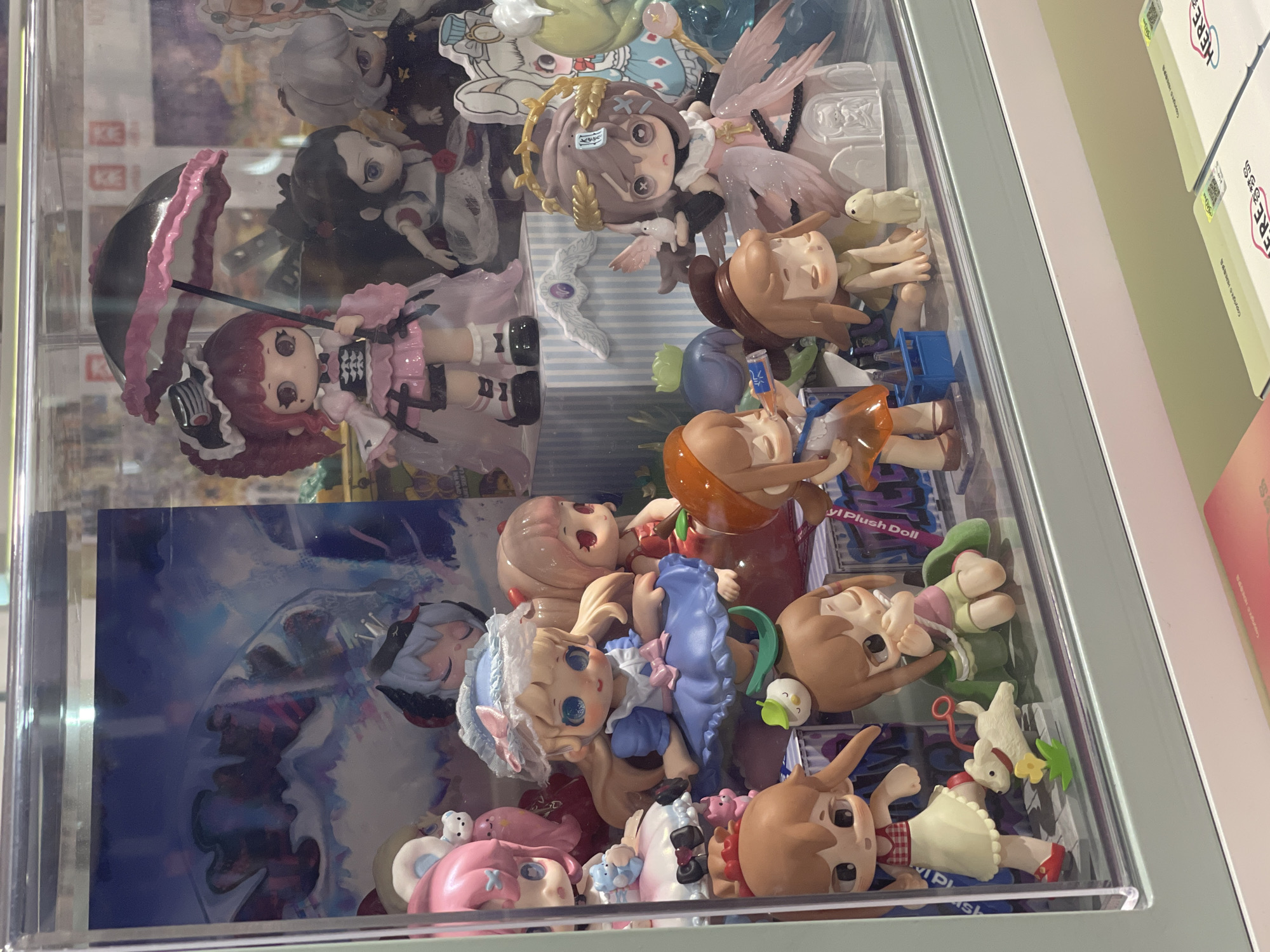 This image shows a display case filled with numerous anime-style dolls. The dolls are in various poses and outfits, with some holding accessories like umbrellas and bottles. The display case itself appears to be made of glass and is illuminated, highlighting the colorful details of the dolls. In the background of the display case, there are printed images that appear to be artwork or scenes related to the dolls. Below the display case, there are some printed materials, possibly packaging or promotional items, with text and QR codes visible. The overall scene suggests a retail environment or a collector's display.