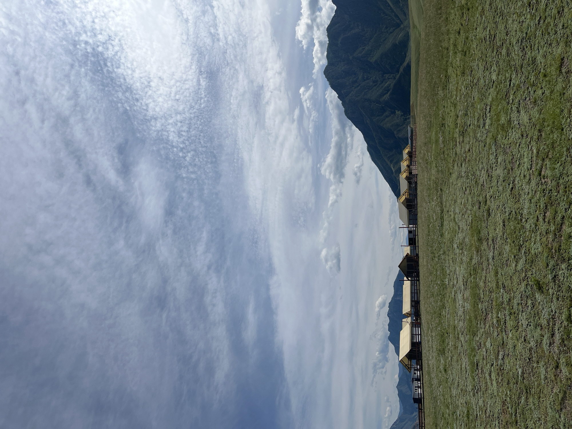 The image shows a vast, green, grassy field under a cloudy sky. In the middle ground, a row of canvas tents with yellow roofs are set up, appearing to be part of a glamping site. A wooden fence runs along the front of the tents. Behind the tents, several dark, rugged mountains rise sharply against the sky, with some smaller mountains visible in the distance. The overall scene suggests a remote, natural landscape. The location is Little Yaloman, Russia.