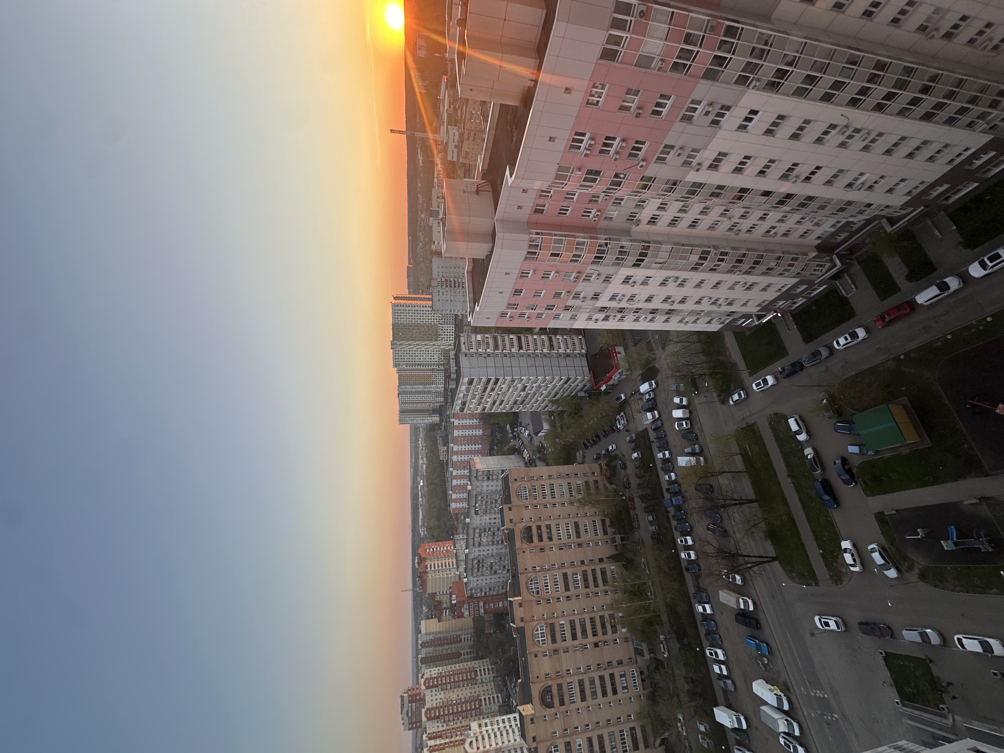 The image is an aerial view of Khimki, Russia, taken at dusk or dawn, with the sun low on the horizon. The city is characterized by a mix of high-rise residential buildings of varying architectural styles. The buildings are mainly constructed of concrete, with some featuring pink and white facades. There are cars parked along the sides of streets. The image shows a relatively high population density.