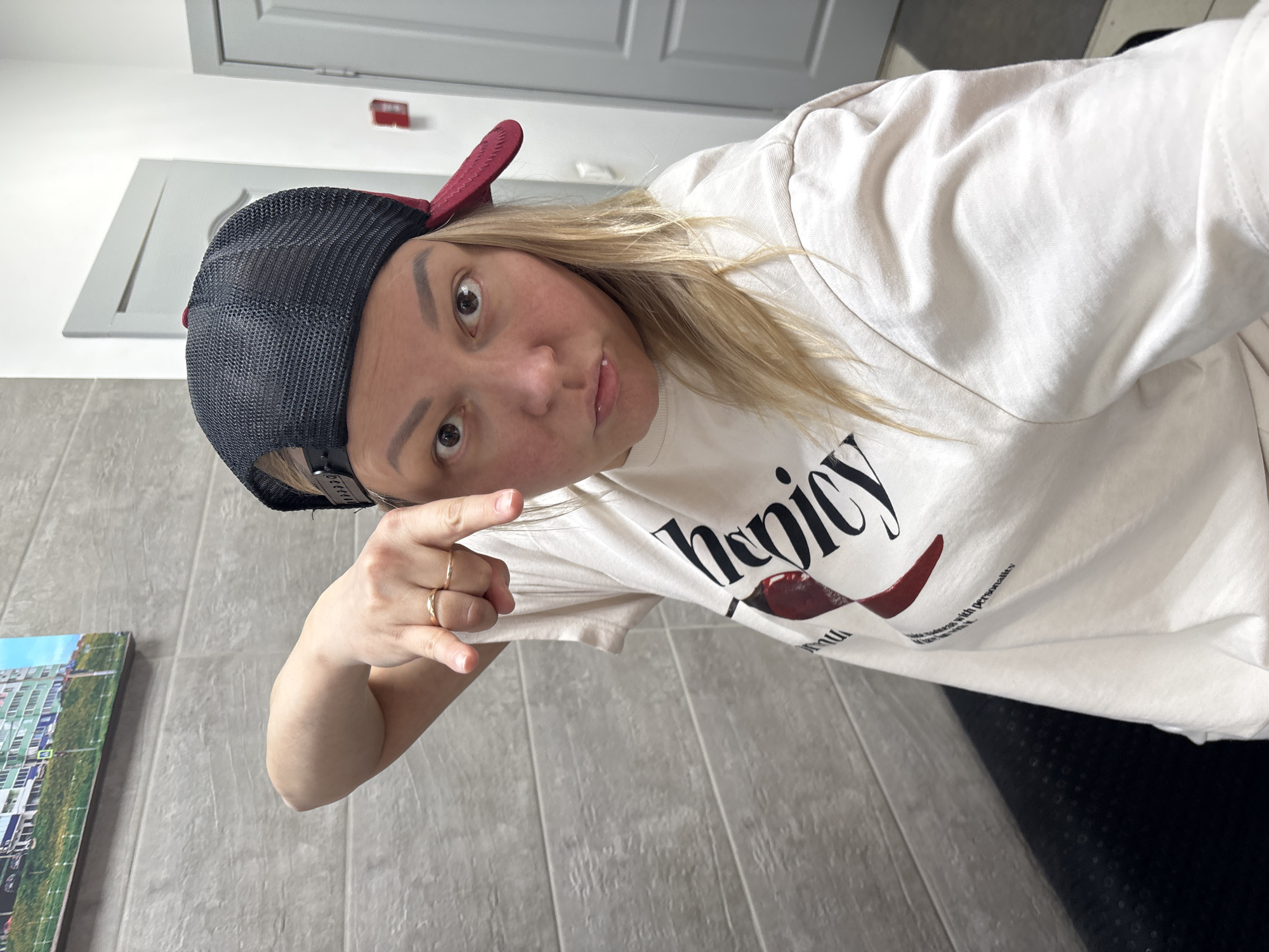 An adult female, with blonde hair and a light complexion, is depicted indoors, taking a selfie. She wears a black and maroon baseball cap backward, a light-colored long-sleeved shirt with "henicy" printed in black and red on the chest, and two gold-colored rings on her left ring finger. Her left arm is bent, and her index finger points downwards. Her eyes are wide, and her lips are pursed in an expressive pose, directly engaging the camera.
The immediate environment consists of a wall tiled with horizontal gray rectangular panels on the left, beneath which a dark, possibly upholstered, surface (likely a bench or couch) is partially visible. A framed picture depicting a light-colored, multi-story residential building on a green slope under a blue sky hangs on the tiled wall. To the right, two light gray or white doors are visible, suggesting a hallway or interior room arrangement. A red rectangular object, consistent with a fire alarm or emergency device, is mounted on the wall between the two doors.
The precise location within the city of Izhevsk, Russia, cannot be determined from the visual information provided. The scene suggests a modern interior setting, possibly a public waiting area or a residential hallway, with architectural elements common to urban environments in Russia.