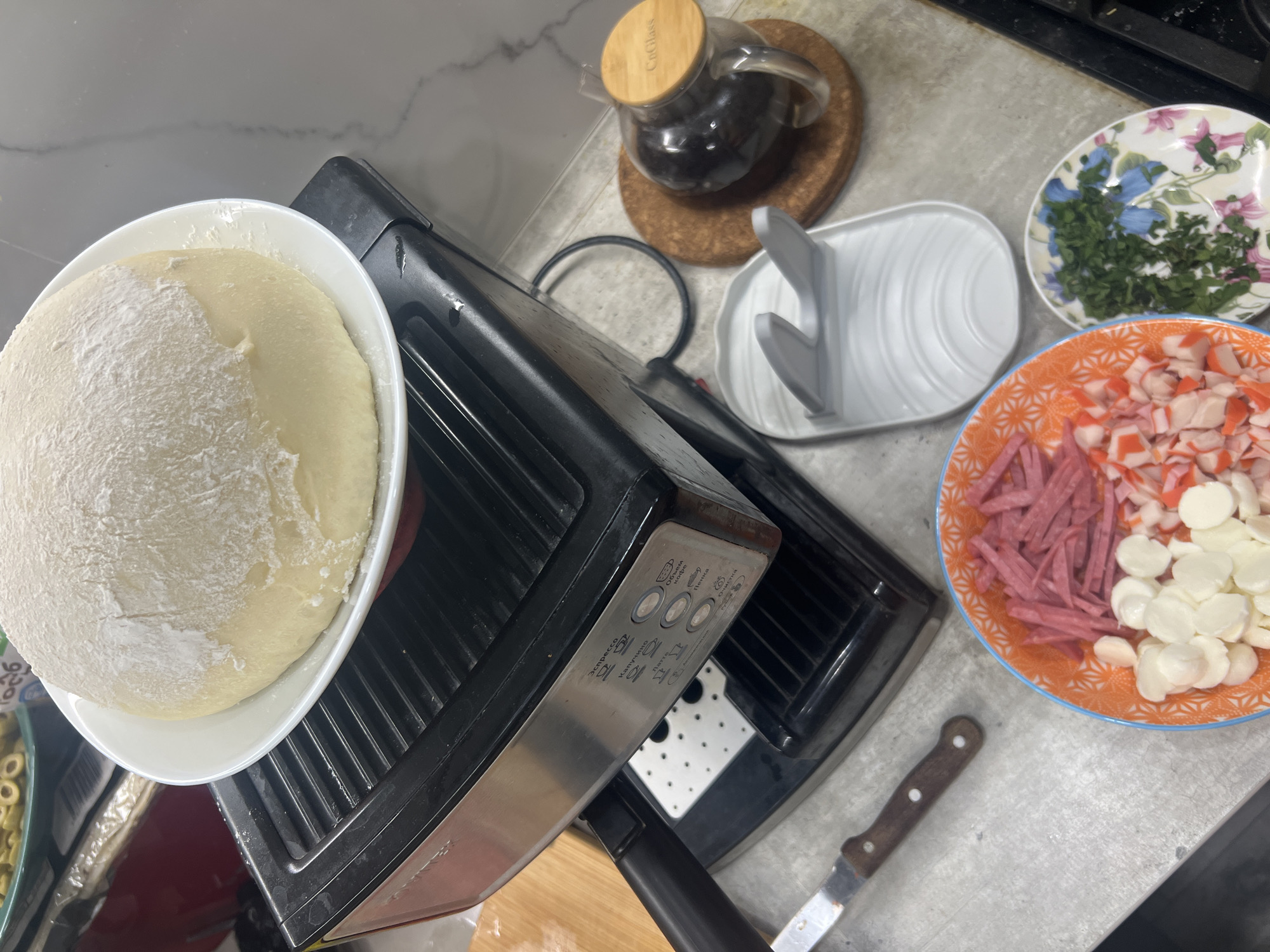 A close-up, overhead shot shows a kitchen counter with various food preparation items and a coffee machine. In the center, a large ball of dough dusted with flour rests on a white plate, which is placed on top of a black espresso machine. To the left of the coffee machine, a wooden cutting board is partially visible. In the foreground, two small plates are arranged. The plate on the left, with an orange and white patterned rim, contains chopped ingredients including what appears to be salami, mozzarella balls, and imitation crab sticks. The plate on the right, with a floral design, holds chopped green herbs. Next to the coffee machine and to the right of the plates is a glass jar with a wooden lid, filled with dark roasted coffee beans, sitting on a cork coaster. A gray spoon rest is also visible on the counter. The background is out of focus, but suggests a domestic kitchen environment. The scene does not provide any information about the city of Volgograd, Russia.