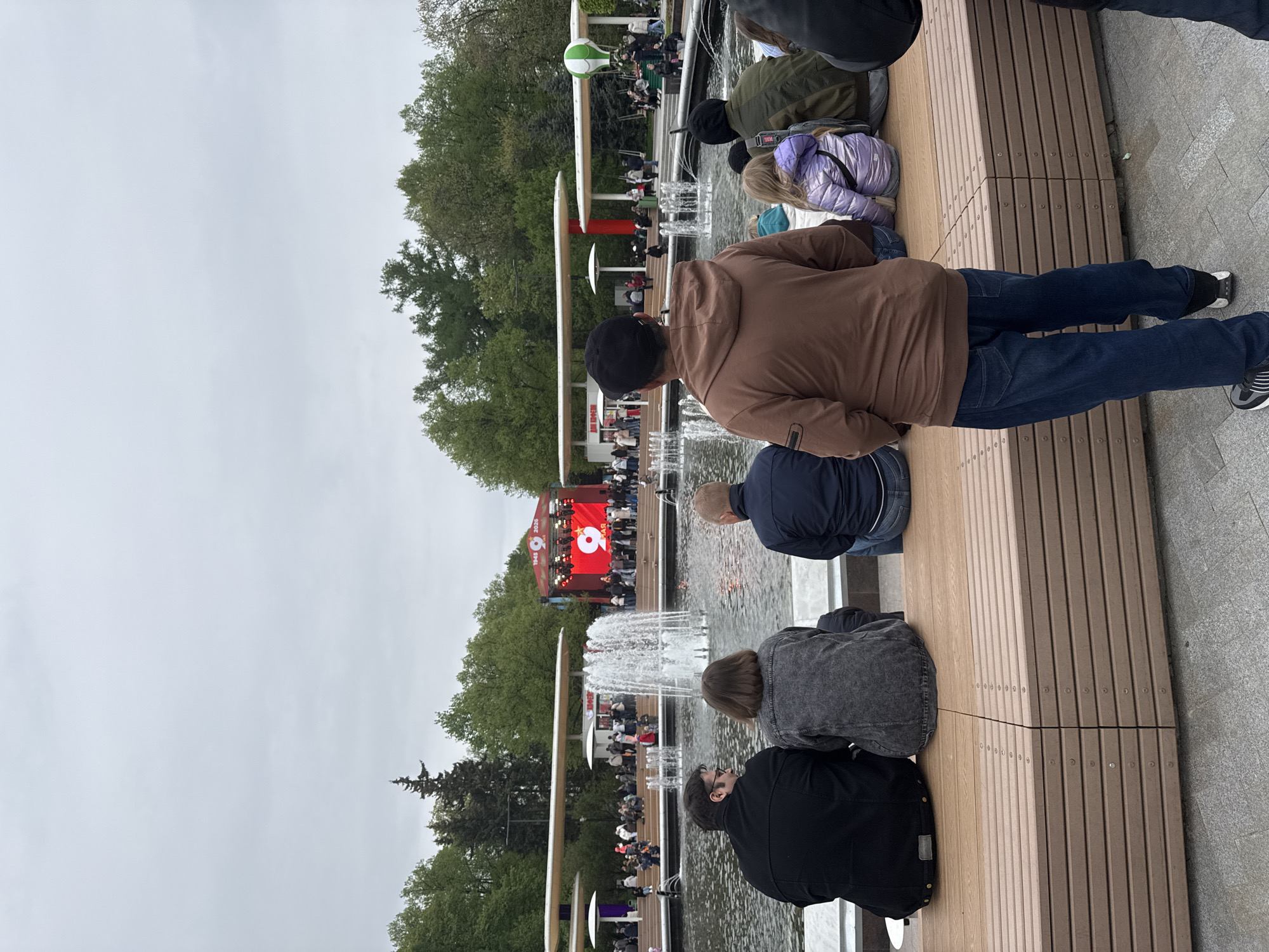 The image shows a group of people sitting on a bench near a fountain in Moscow, Russia. In the background, a large stage with a screen is visible, showing a red display with the number "9" and the dates "1945" and "2026." People are gathered around the stage, suggesting an event or performance. The weather appears overcast. The bench is made of wooden planks, and the fountain has multiple jets of water. There are trees and buildings in the distance, indicating a park-like setting.