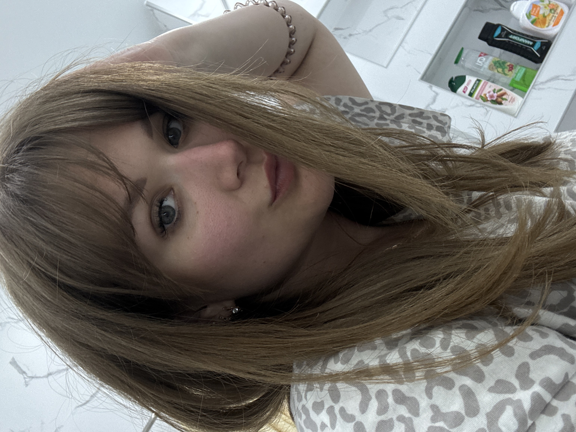 A person with long, light brown hair, bangs, and blue eyes is posing for a selfie. The individual is wearing a bracelet and is dressed in a patterned shirt. In the background, there's a shelf with toiletries. The location is possibly a bathroom. There is no information provided to confirm that this image was taken in Stavropol, Russia.
