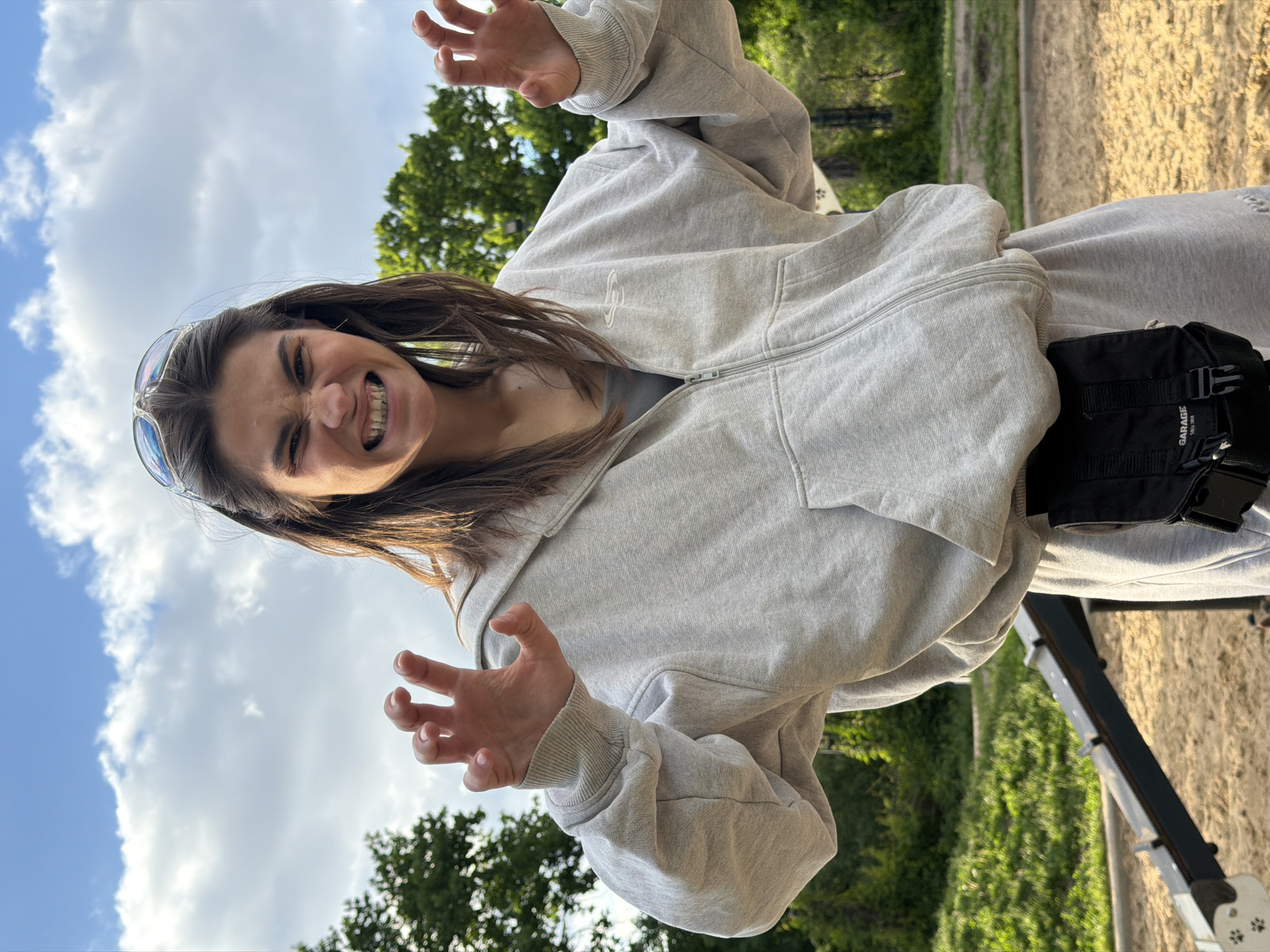 A young woman stands outdoors, likely in a park, given the greenery in the background and visible trees. She wears a light grey zip-up hoodie and matching sweatpants, along with sunglasses perched on her head. A black bag is secured around her waist. She is making a playful, clawing gesture with both hands, and her facial expression shows teeth bared in a playful grimace. The sky is visible, partially cloudy. A portion of what appears to be playground equipment is also visible. There is no definitive information to confirm location in Moscow, Russia, or any specific city.