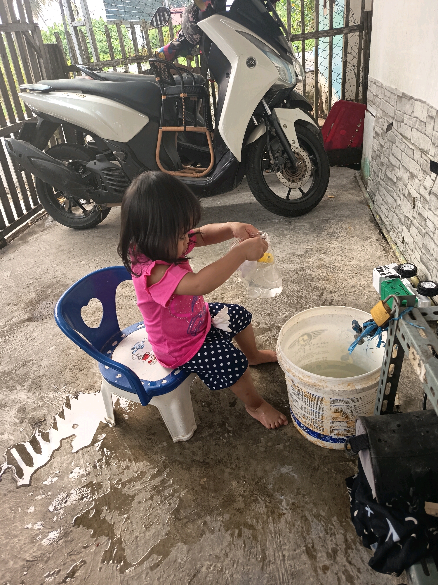 A young girl sits on a small blue and white plastic chair in what appears to be an outdoor covered area, possibly a carport or patio, in Banjarmasin, Indonesia. She is wearing a pink t-shirt with cut-out shoulders and navy blue polka dot capri pants. In her hands, she holds a clear plastic bottle filled with water and a yellow object, seemingly playing with it. The ground is wet, with puddles of water reflecting the light, suggesting recent activity or a spill. To the left of the child, a white and black motorcycle is parked. A large white bucket filled with water is visible to the right of the girl. In the background, a metal gate and a brick wall can be seen. The overall scene depicts a child engaged in play in a somewhat cluttered outdoor domestic environment.