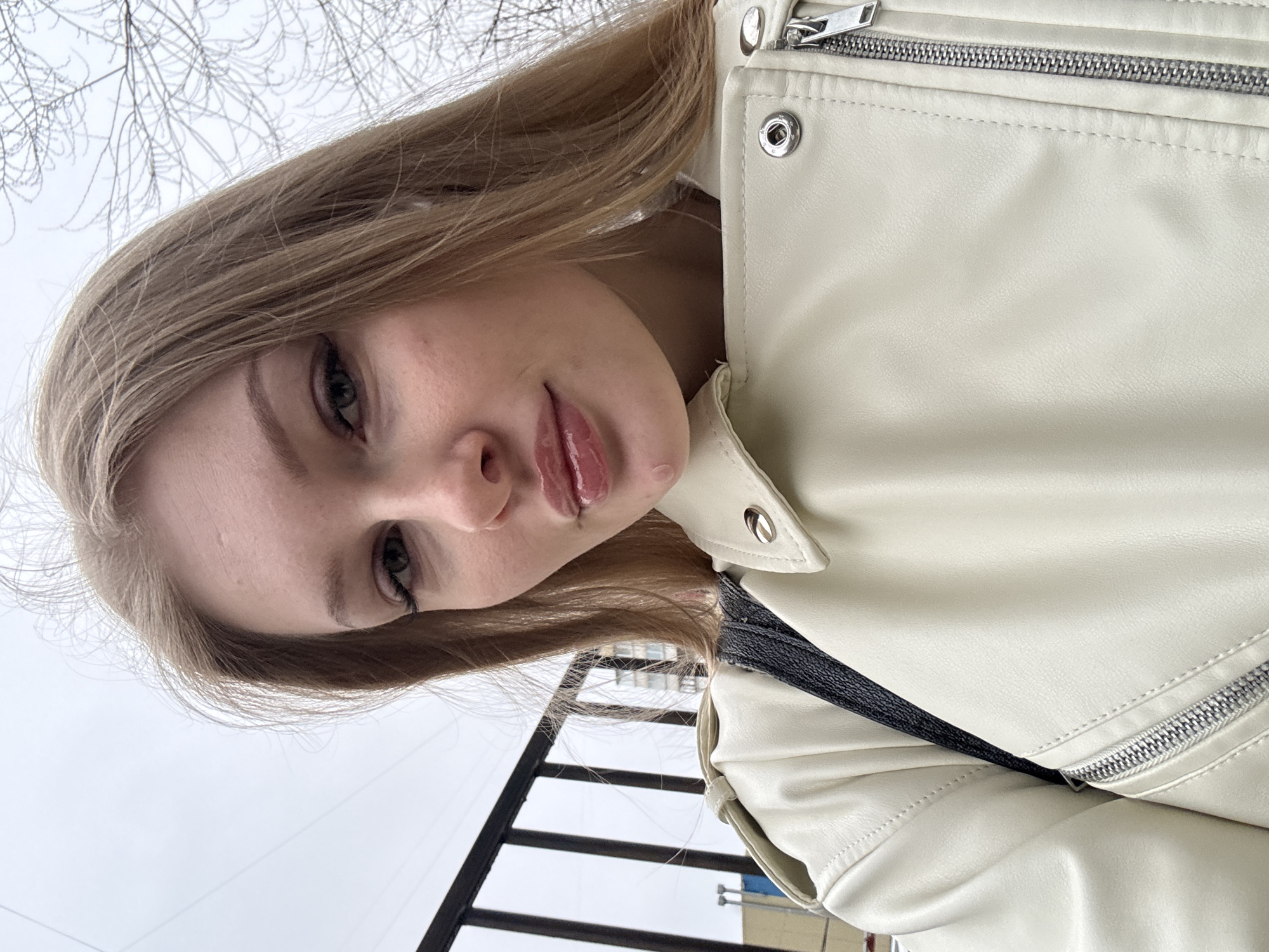 A fair-skinned young woman with long blonde hair, wearing a white leather jacket, is photographed from a low angle, suggesting the camera is below her. The background includes a gray sky and bare tree branches, indicating a possible late autumn or winter setting. A black metal railing is visible near the bottom of the frame. The woman is likely in Perm, Russia.