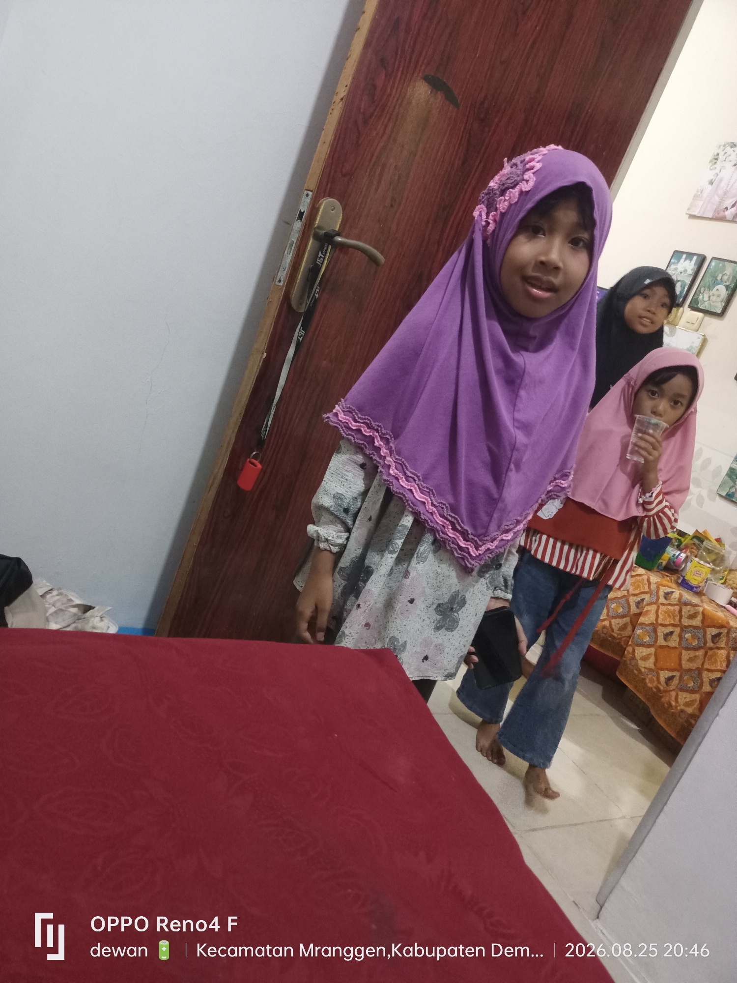 The image shows three young girls in a room. The girl in the foreground, on the left, is wearing a purple hijab and a floral-patterned shirt. She is looking at the camera. To her right, another girl is wearing a pink hijab and a striped shirt, holding a clear plastic cup. Behind them, partially visible, is a third girl wearing a black hijab. The background features a wooden door with a handle, a white wall, and a table covered with a patterned cloth, holding various items including snacks. The bottom of the image displays a watermark indicating "OPPO Reno4 F dewan | Kecamatan Mranggen, Kabupaten Dem... | 2026.08.25 20:46". The setting appears to be indoors, likely a residential space in Mranggen, Indonesia, given the watermark.