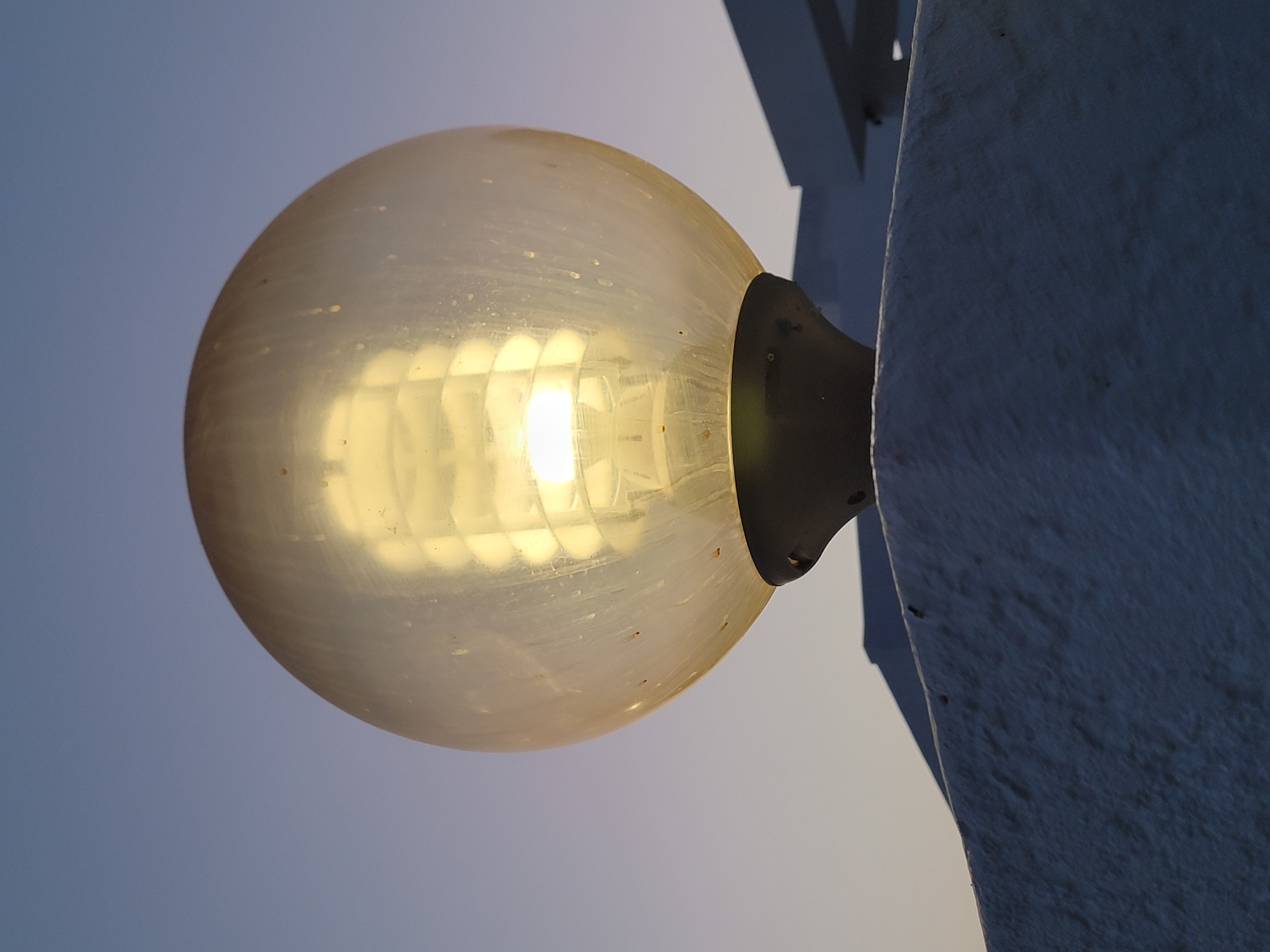 A close-up, low-angle shot captures a lit, spherical outdoor lamp against a dusk sky. The lamp has a translucent yellow-orange globe, which appears slightly dirty, revealing a spiral fluorescent bulb inside that emits a bright, warm light. The globe is mounted on a dark, conical base. In the background, parts of a white, modern building with a flat roof and horizontal architectural elements are visible, suggesting an urban setting. The sky transitions from a pale blue at the top to a lighter, almost white hue near the horizon, indicating the end of daylight. The foreground shows a textured, white concrete surface, possibly a wall or pillar, with a rough, stucco-like finish. The overall scene is static and devoid of any human presence. The image was taken in Quarteira, Portugal.