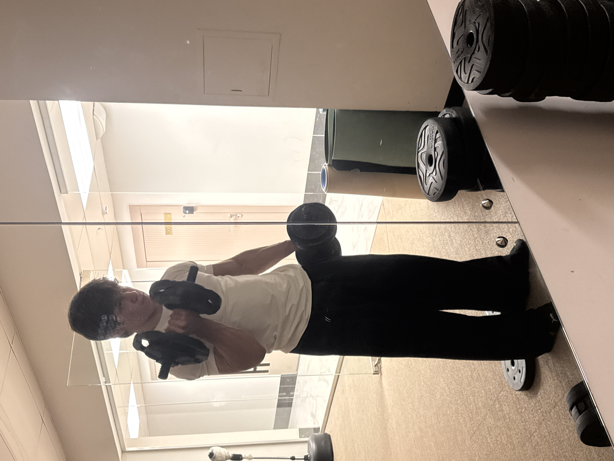 A young man, likely in his late teens or early twenties, is depicted in his mirror reflection while exercising in an indoor fitness area. He stands upright, wearing a white t-shirt and black pants, engaged in bicep curls with two black dumbbells. His expression is focused and neutral as he observes his form in the mirror.
The setting appears to be a well-lit gym or fitness corner, potentially within a hotel or residential complex in Khimki, Russia. The room features light-colored walls and a light beige or brown carpeted floor. A prominent mirror occupies the foreground, reflecting the man and elements behind the camera's position.
Visible objects include the two black dumbbells being used, and a stack of similar weight plates or dumbbells on a rack to the right. Another single weight plate lies on the floor near the man's feet. Reflected in the background are an interior door with the number "800" visible, suggesting a room number, and a white panel or vent on the wall above the mirror. A green mat or rolled-up item is also discernible in the reflection. The brand name "ATTAIN" is visible on the textured ends of the dumbbells. The lighting is bright, indicating an illuminated indoor environment.