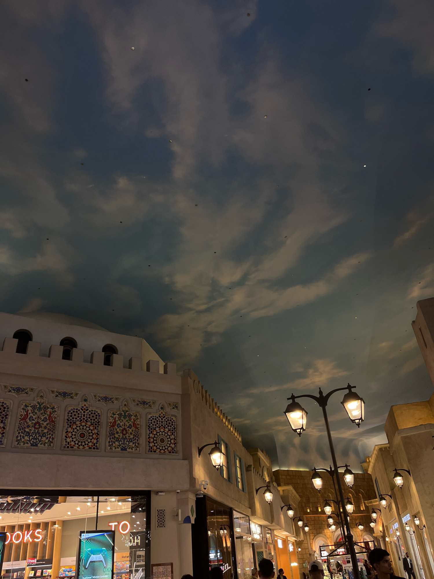 This image captures a nighttime scene within a shopping arcade in Dubai, United Arab Emirates, designed to resemble an old-world streetscape with an artificial sky ceiling. The architecture features buildings with intricate decorative tiling and crenellated parapets, evoking a Middle Eastern aesthetic. Ornate lampposts with multiple lights illuminate the walkway, casting a warm glow. In the foreground, a shop window displays merchandise, with neon lettering partially visible, reading "BOOKS" and "TO". People are visible in the lower portion of the image, walking through the arcade. The ceiling is a painted panorama of clouds, creating the illusion of an open sky. The overall impression is one of a meticulously crafted indoor environment designed for a unique shopping and entertainment experience.