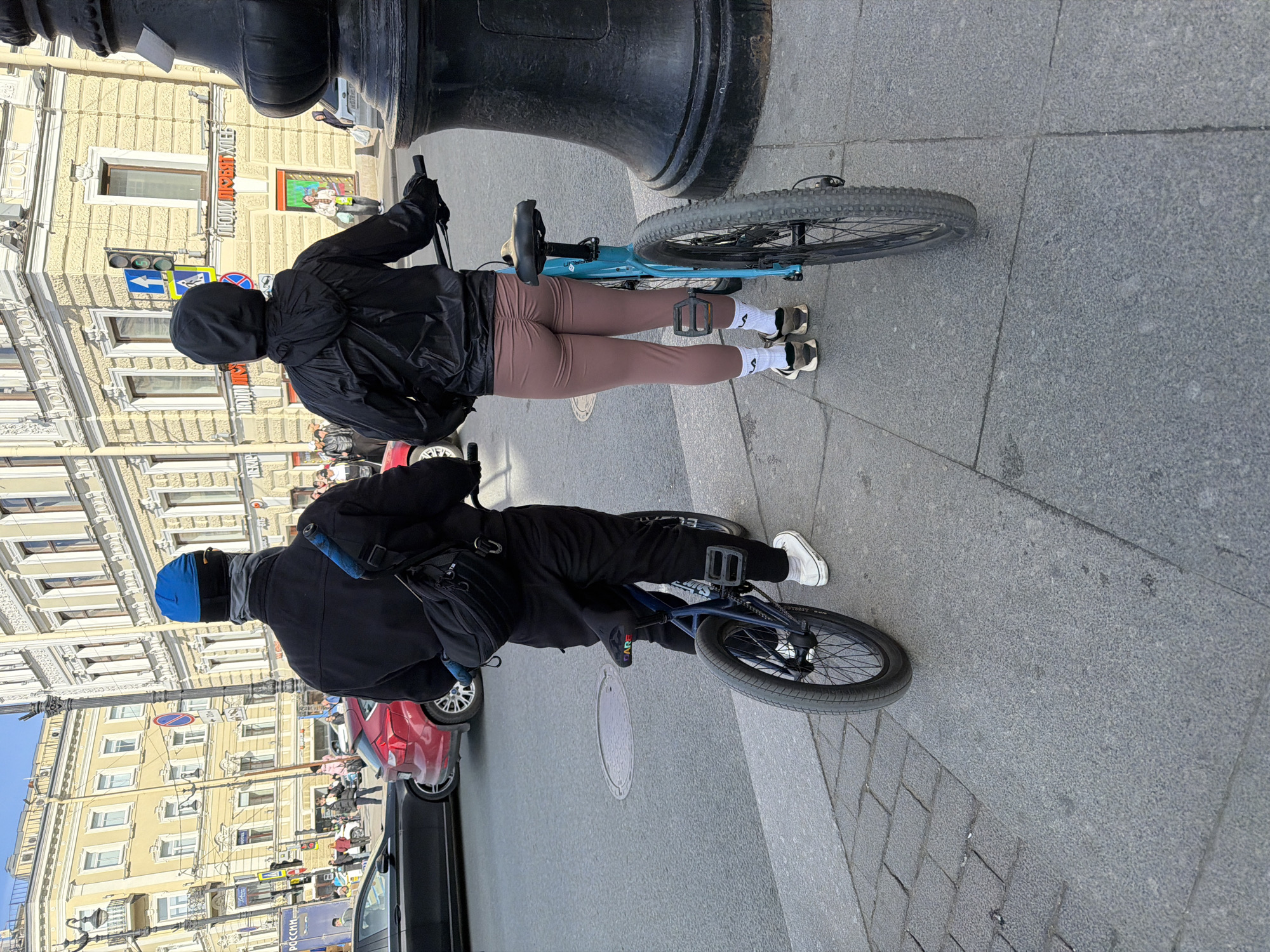 Two people on bicycles are standing on a sidewalk in Saint Petersburg, Russia. The person closer to the camera is wearing a blue balaclava, a black jacket, and black pants, and is riding a blue bicycle. The other person is wearing a black hooded jacket and brown leggings, and is also riding a blue bicycle. They are both facing away from the camera. Behind them is a street with cars and buildings. The buildings are light yellow with ornate architectural details. There are traffic lights and street signs visible. A street lamp post is in the foreground.