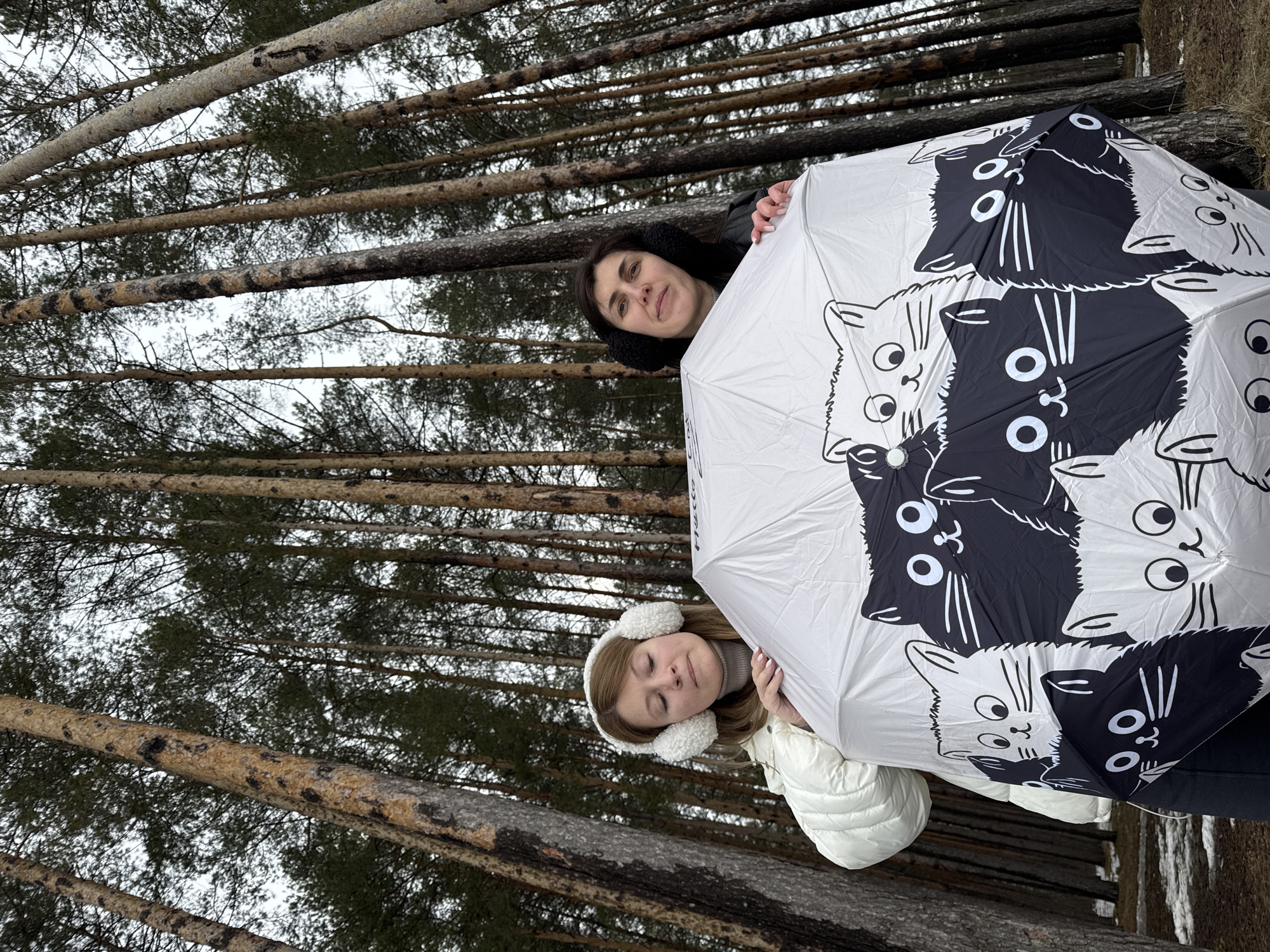 Two women are standing behind a large white umbrella with alternating black and white cat faces printed on it. The woman at the top of the frame has dark hair and is wearing black earmuffs and a black jacket. The woman at the bottom has blonde hair and is wearing white earmuffs and a white puffer jacket. They are standing in a forest of tall, slender trees with light-colored trunks and evergreen foliage. The sky is overcast. This scene is in the city of Chaikovsky, Russia.