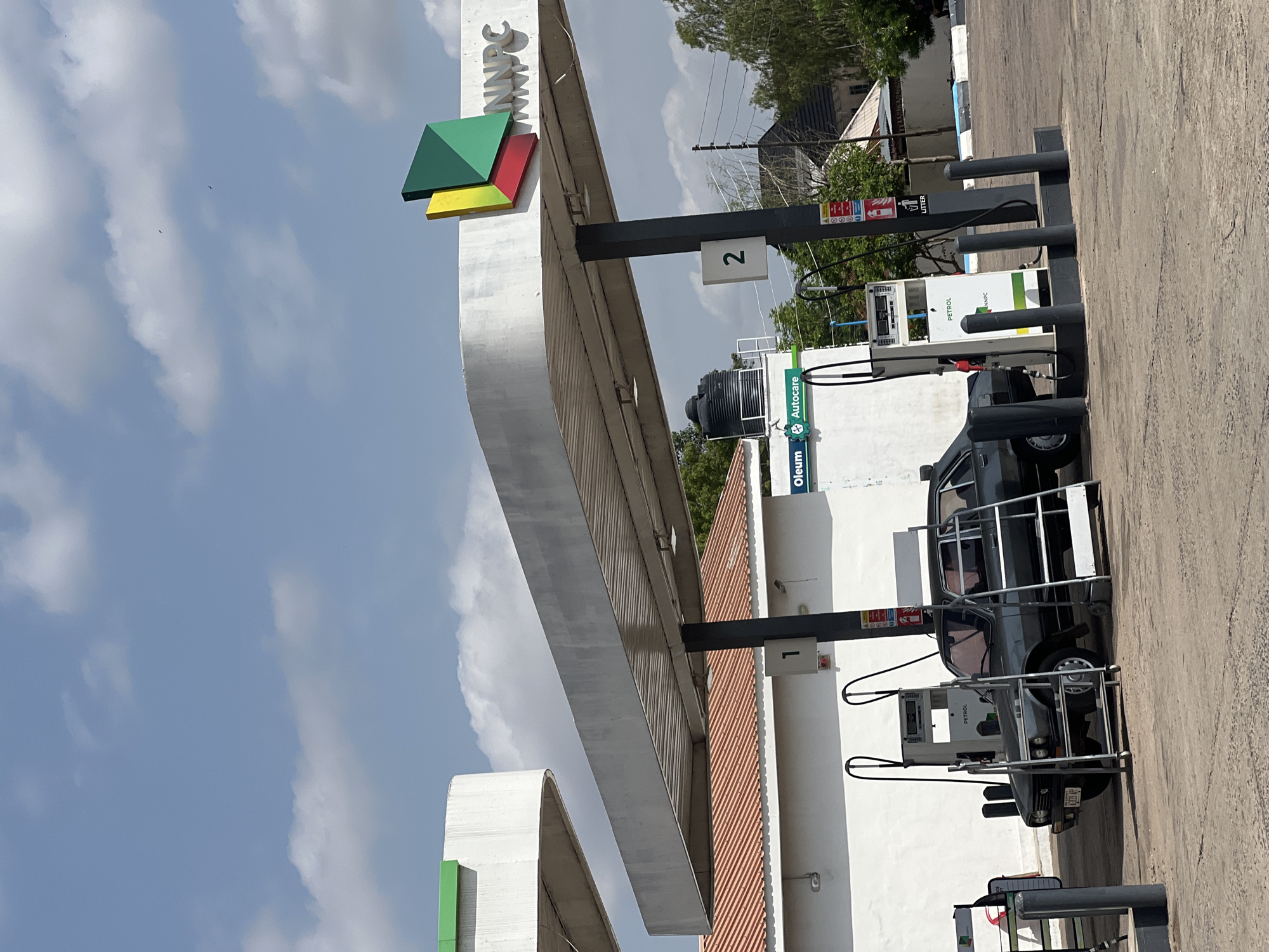 The image shows a scene at an NNPC gas station in Jalingo, Nigeria. A dark grey sedan is parked next to a petrol pump. The car has a license plate visible. Above the pump, there is a sign that says "PETROL" and "NNPC". A black water tank is mounted on the roof of a white building behind the gas station. The gas station has a large canopy supported by pillars. A sign with the NNPC logo, featuring three colored geometric shapes, is visible on the canopy. The sky is blue with scattered white clouds. The ground is paved. There are no people visible in the image.