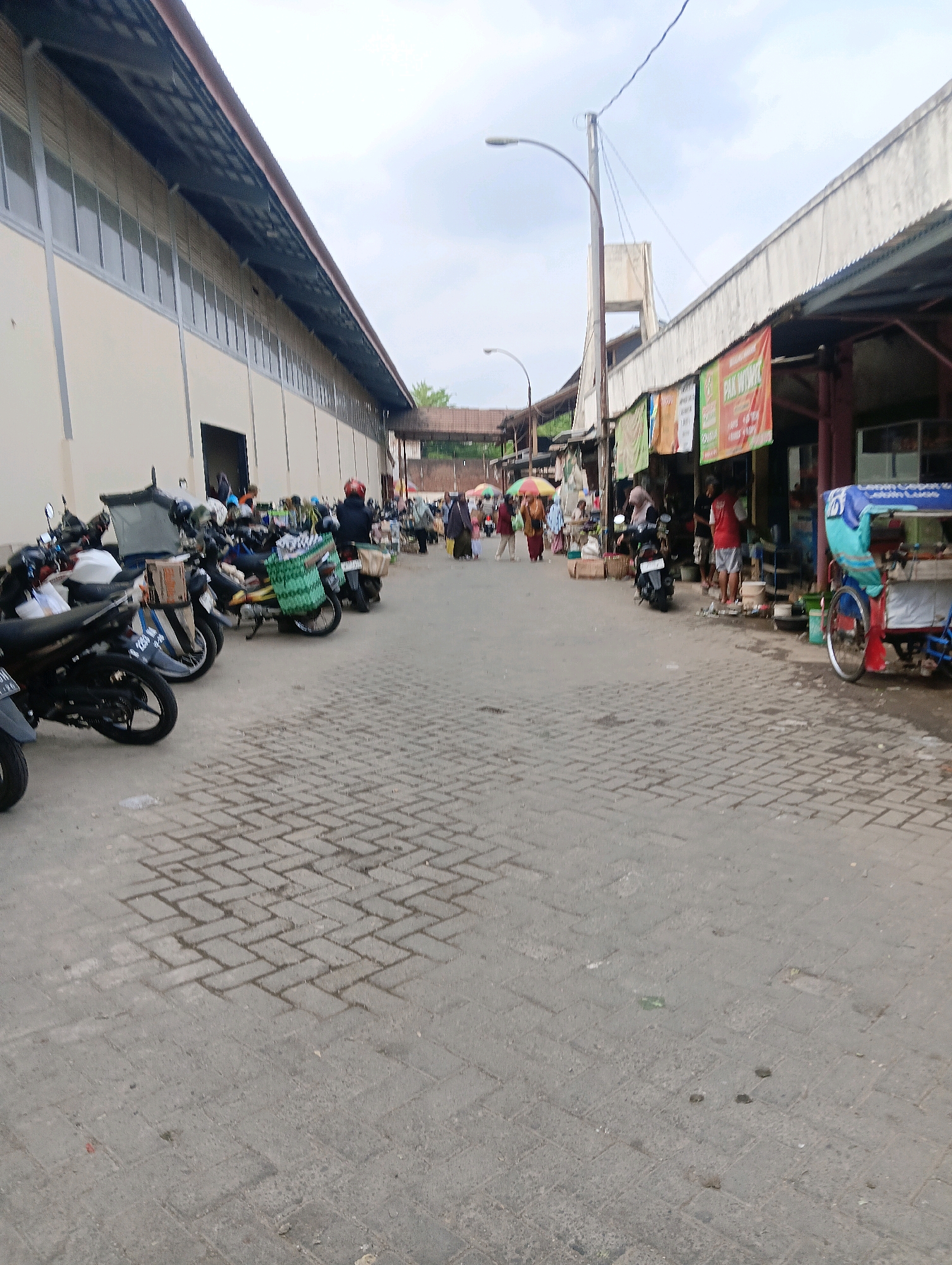 This is a street scene in Sokaraja, Indonesia. On the left is a long, light-colored building with a dark, corrugated metal roof. The building has a row of windows along its upper portion. A line of motorcycles is parked along the sidewalk in front of this building. On the right side of the street, there are various stalls and shops with awnings and hanging banners. Several people are walking and standing in the street, and a few motorcycles are also present. A small, colorful motorized cart, possibly a food vendor, is parked on the right side of the street. The ground is paved with interlocking bricks. The sky is overcast.