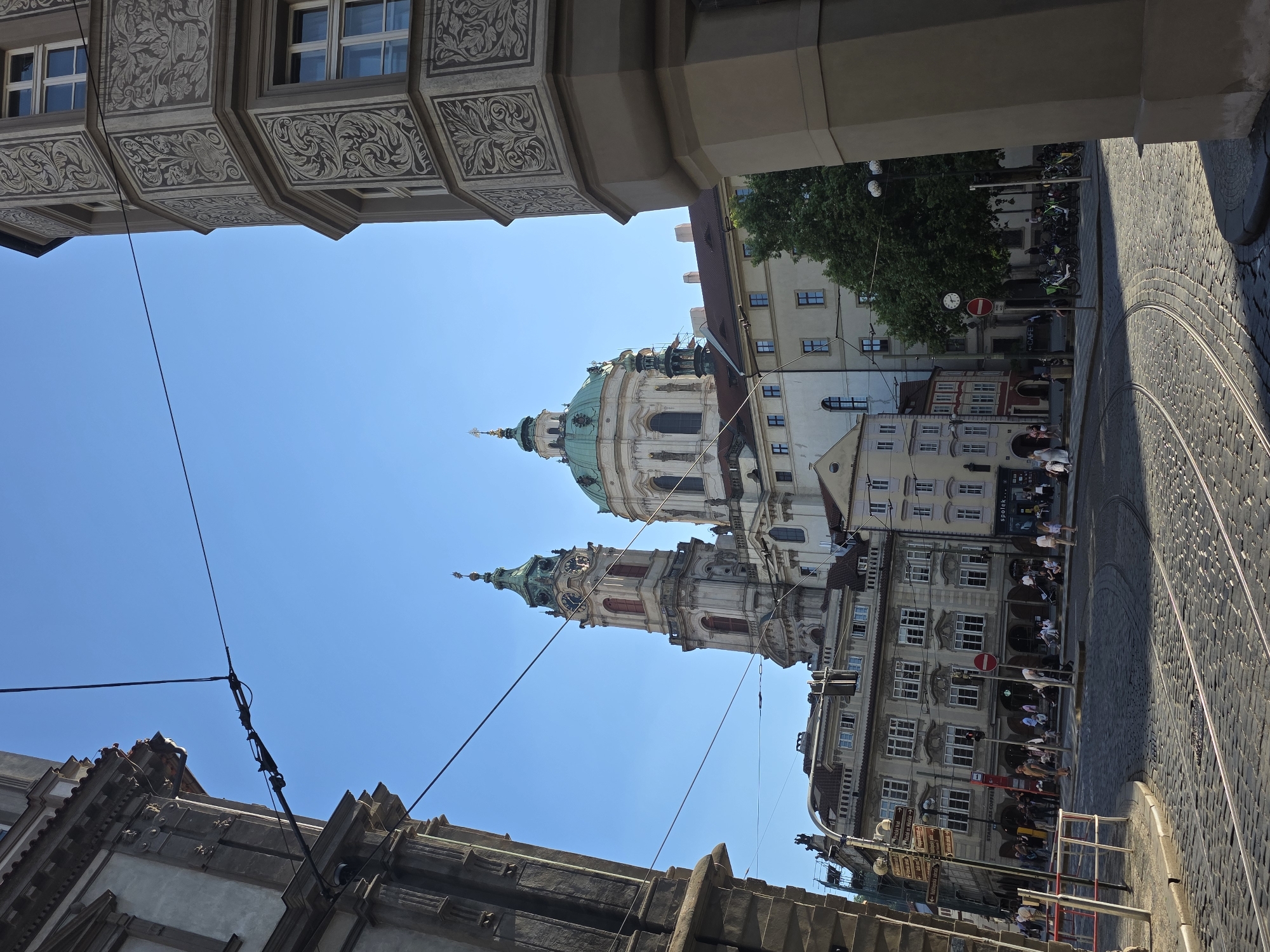 The image captures a view of St. Nicholas Church in Prague, Czech Republic, framed by surrounding buildings. The church's dome and tower are visible against a clear blue sky. The buildings framing the church exhibit ornate architectural details. A section of tram tracks runs along the right side of the image on a cobblestone street. People are seen walking on the sidewalk next to the buildings. Overhead tram lines are also visible.