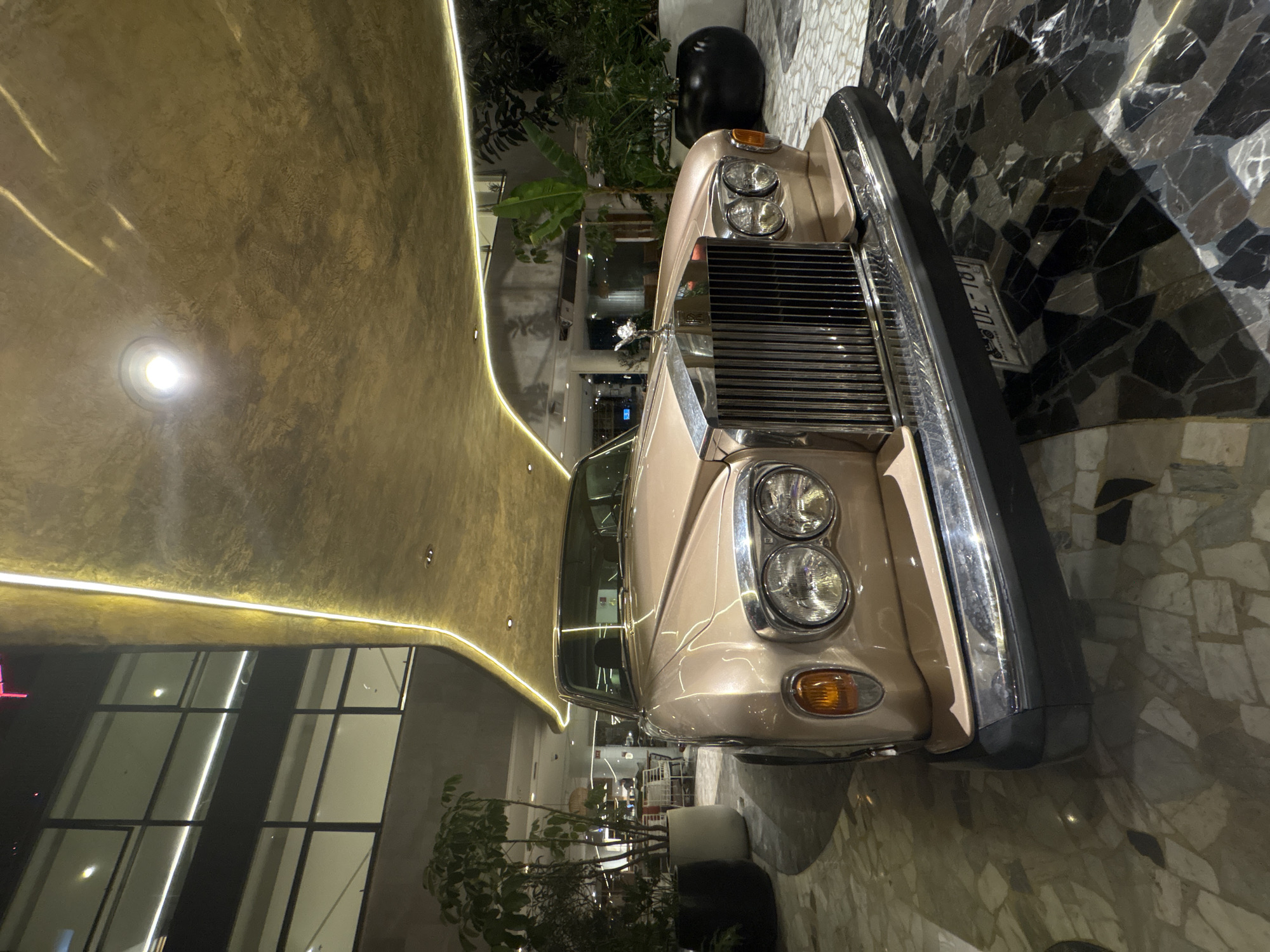 The image shows a cream-colored classic Rolls-Royce car parked inside what appears to be a commercial space. The environment includes a ceiling with integrated lighting, large glass windows, and decorative plants. The floor has a patterned marble or stone surface. In the background, there are tables and chairs, suggesting a restaurant or cafe setting. The car is the primary focus, positioned prominently within the space. The location is likely in Mexico City, Mexico.